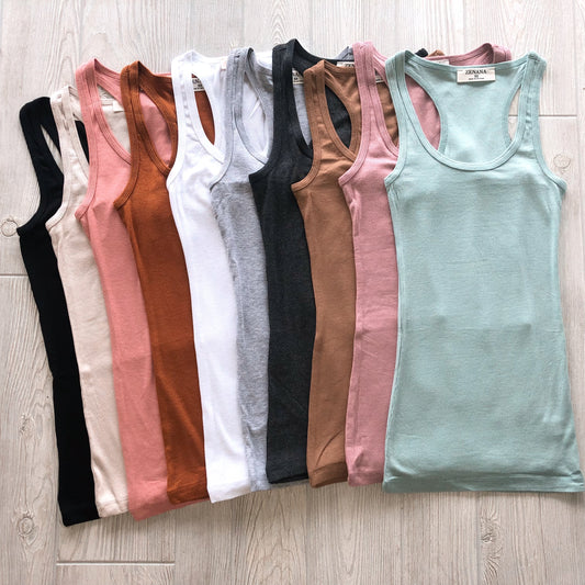 Nine New Basic Racerback Tank tops in colors like black, cream, blush pink, rust, white, gray, brown, tan, and light green are neatly arranged side by side on a light wooden floor. More colors available!.