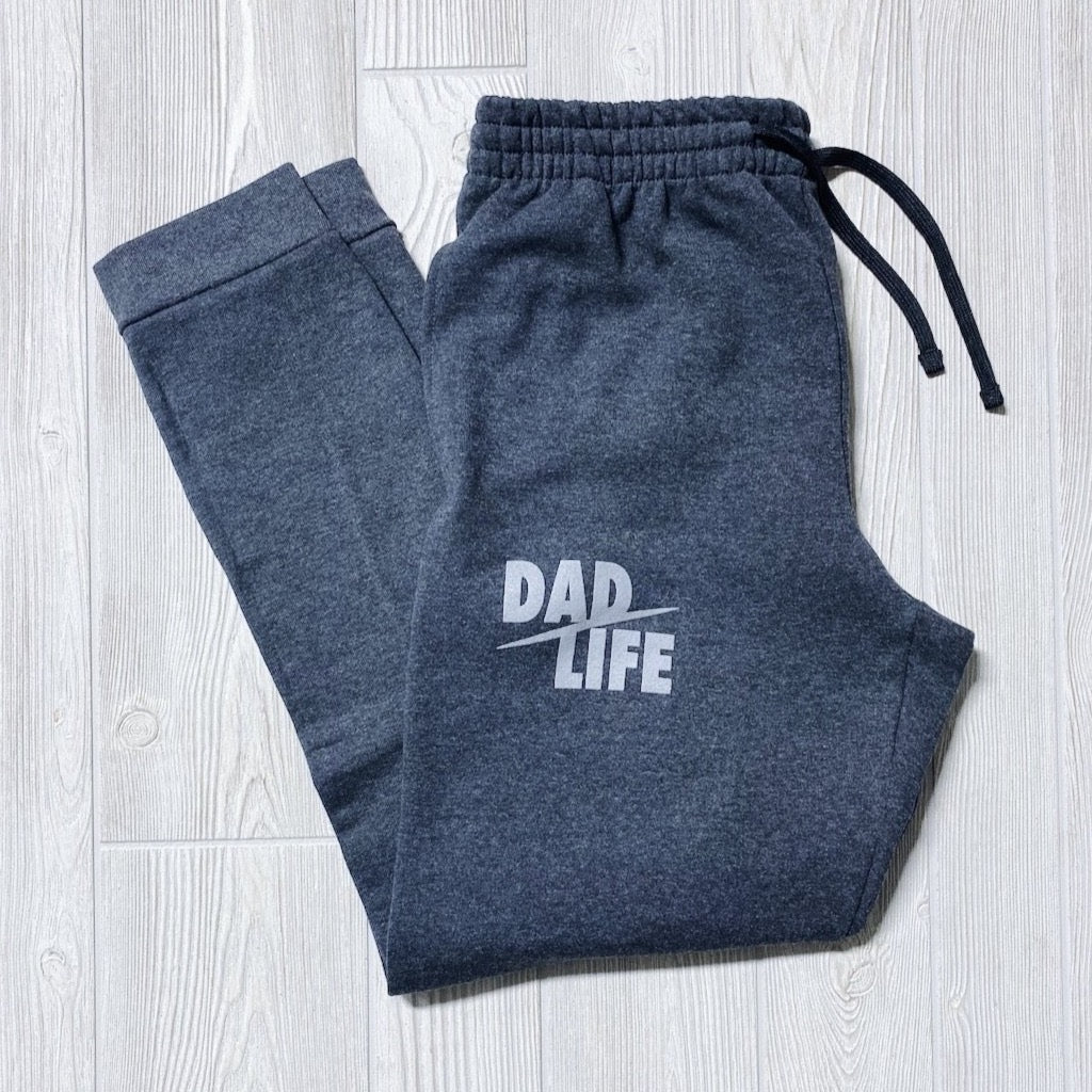 A pair of Dad Life Joggers in charcoal, featuring DAD LIFE in bold white print on the left thigh, displayed flat on a light wood floor.