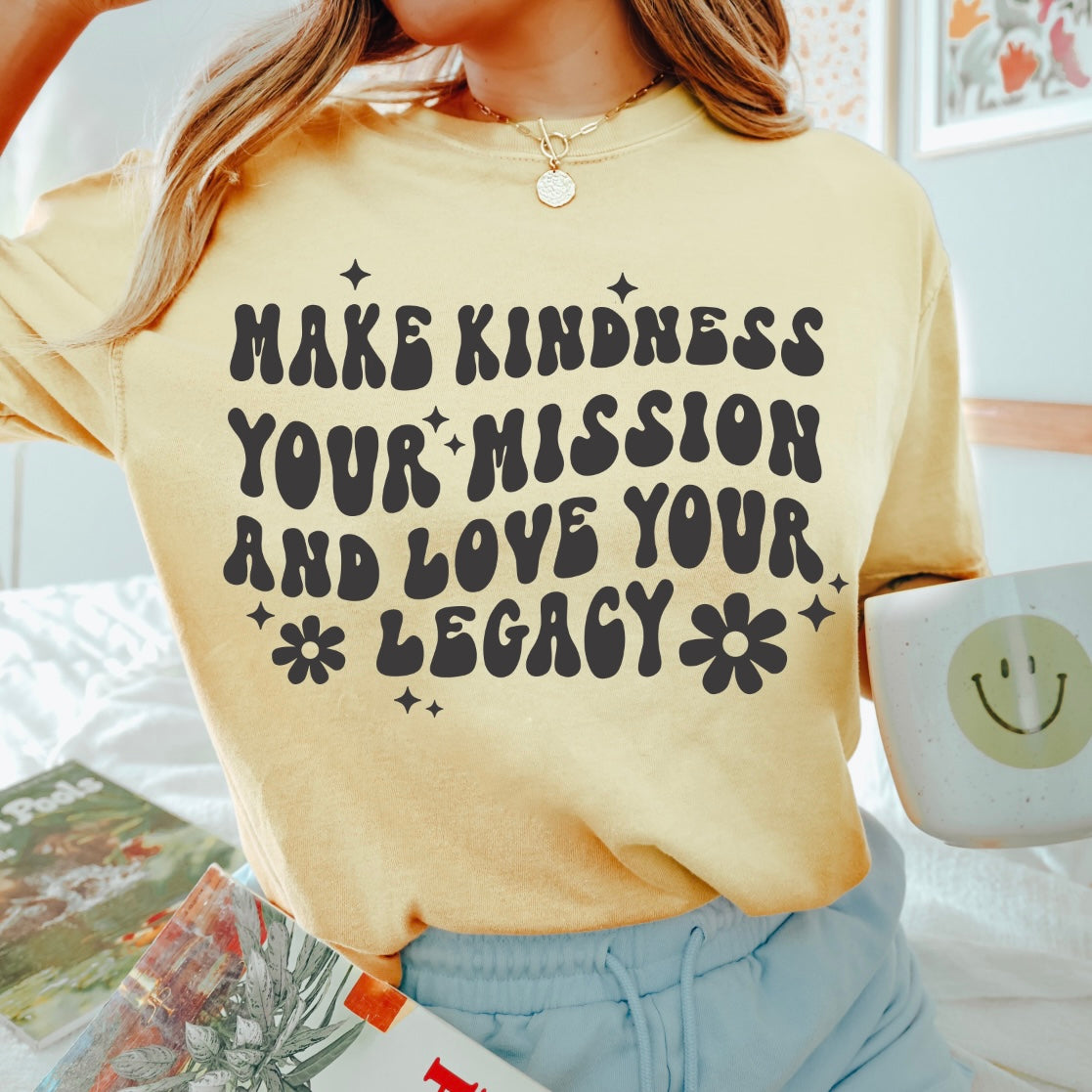 A person wearing the yellow Legacy • Comfort Colors Tee reading Make kindness your mission and love your legacy, holds a white mug with a green smiley face; magazines are on the bed in the background.