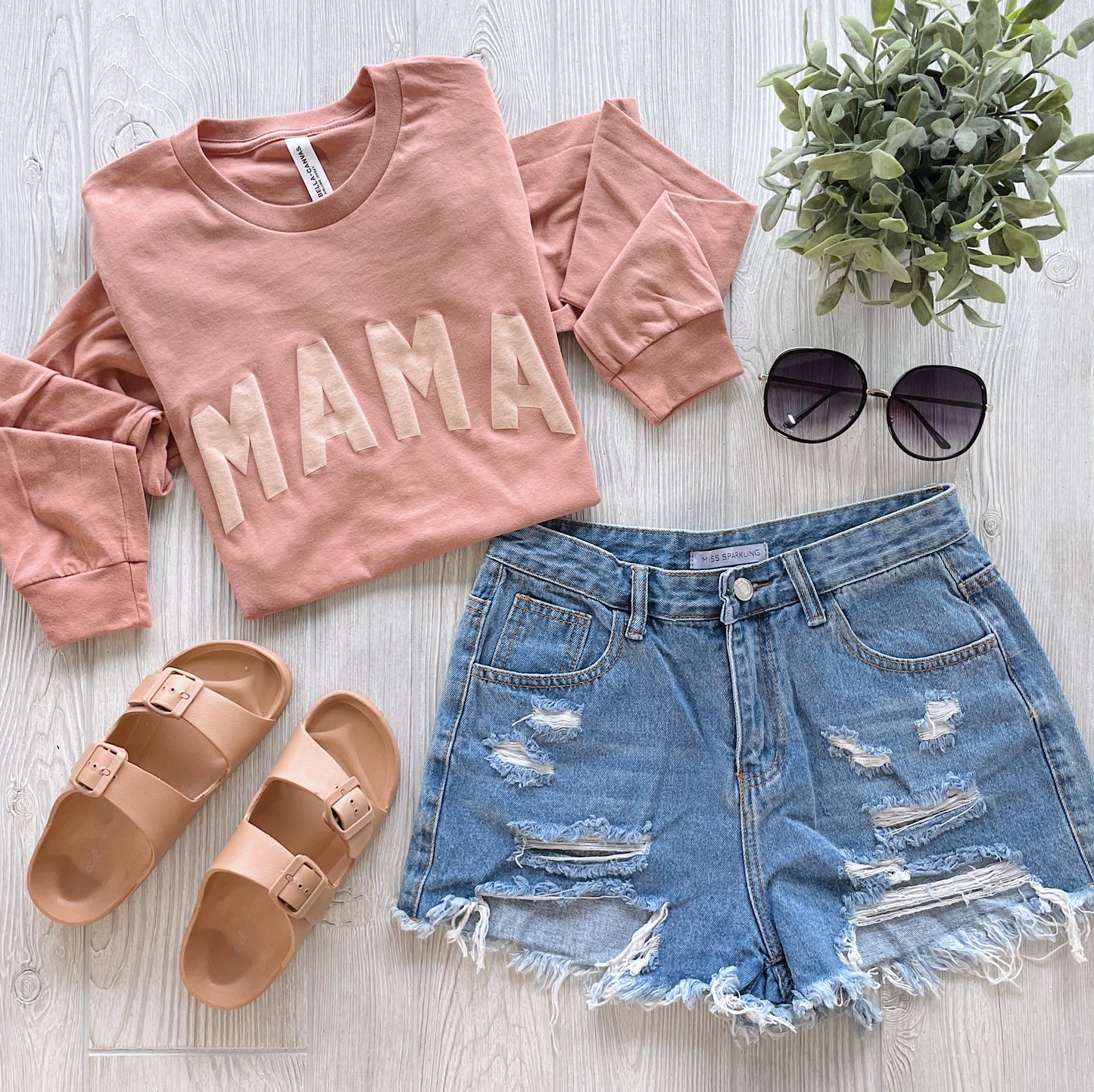 A flat lay of the MAMA • Terracotta Long Sleeve sweatshirt with distressed denim shorts, tan sandals, black sunglasses, and a small potted plant on a light wooden surface.