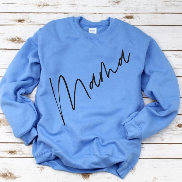 The New Mama Script • Columbia Blue Pullover features diagonal bold black Mama Script text on the front, shown on a white wooden surface.