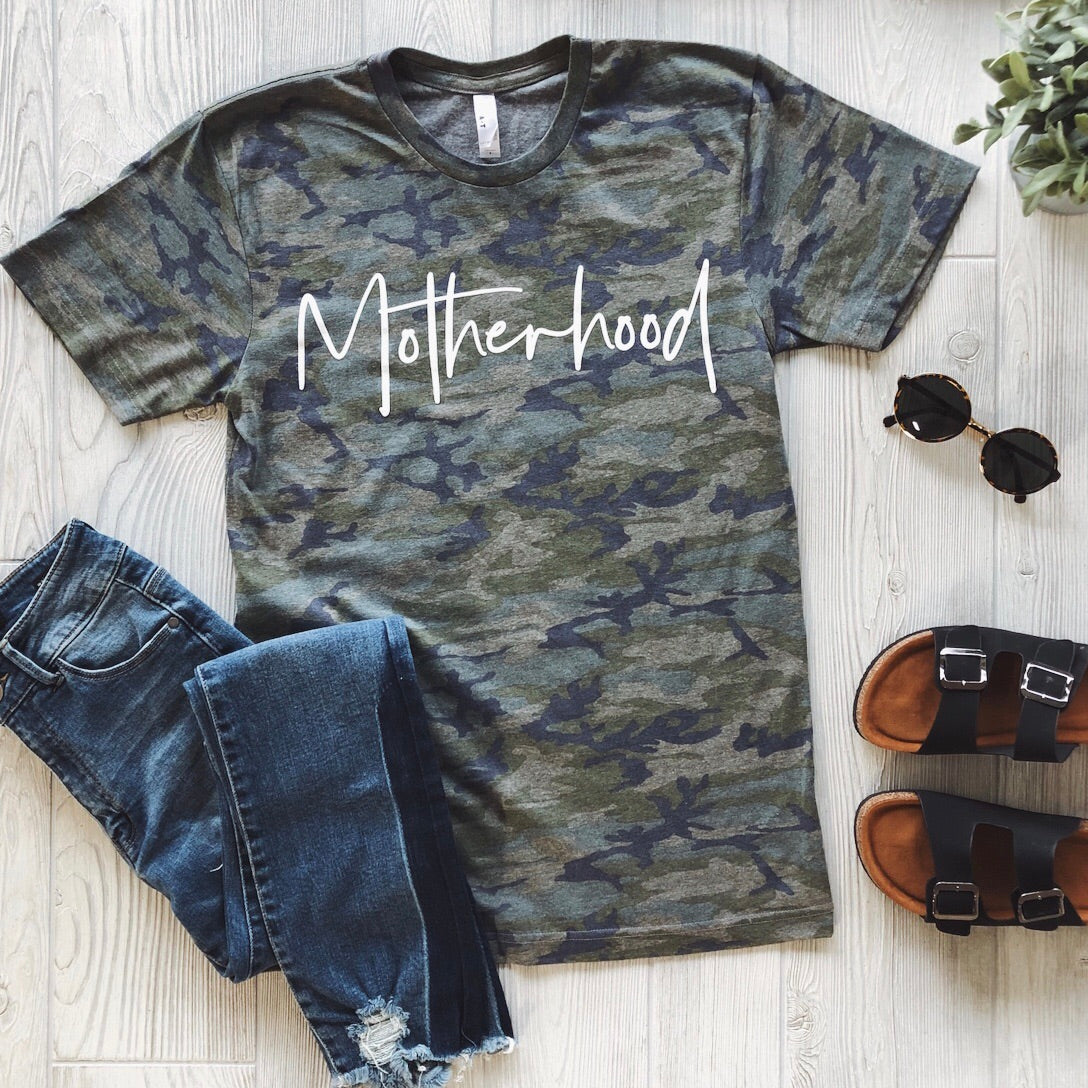 The Motherhood • Vintage Camo Tee features bold white script, styled with blue jeans, black sandals, sunglasses, and a small potted plant on a light wood floor; unisex sizing ensures the perfect fit.