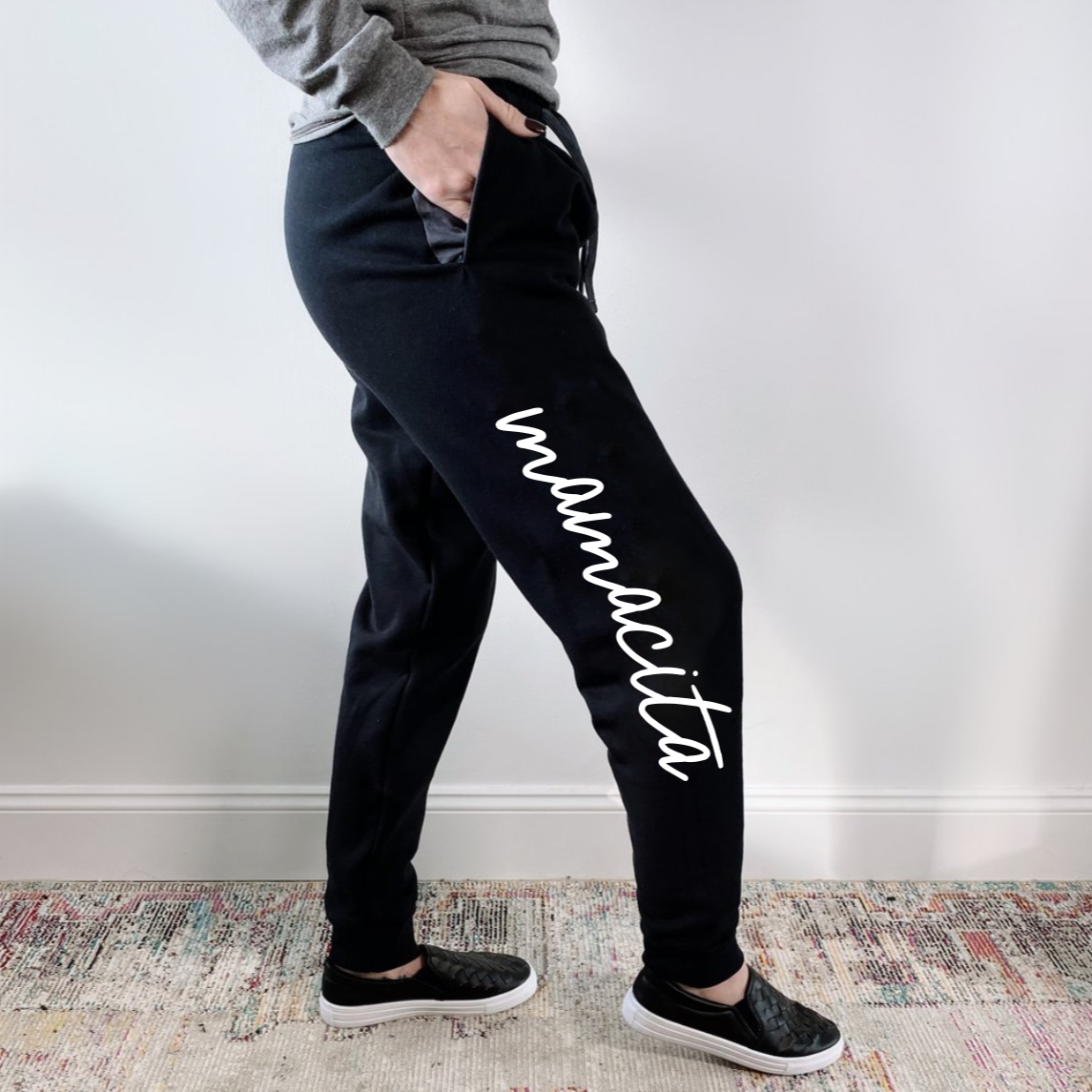 A person wears Mamacita Joggers • black, featuring mamacita in white script down the leg, standing sideways on a patterned rug—a stylish and cozy loungewear essential.