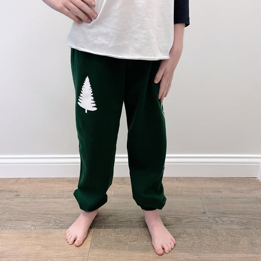 A child’s bare feet stand on a wooden floor, showing only the lower half in dark green Fresh Sweatpants • Kids with a white pine tree graphic on one leg and a white shirt.
