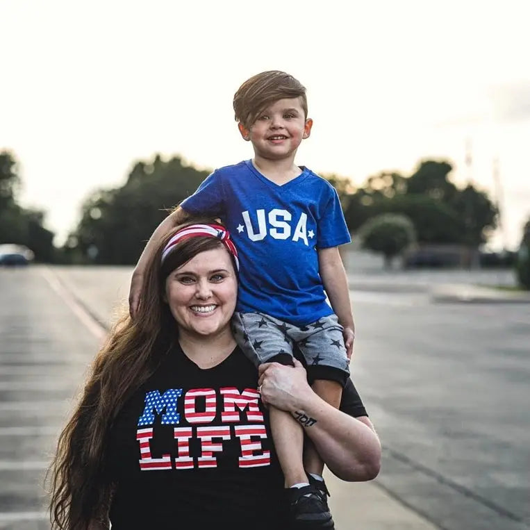 A smiling woman wears the Mom Flag Print Black Tee (unisex fit) and carries a boy in a USA shirt on her shoulders, celebrating Americana style outdoors in a tree-lined parking lot.