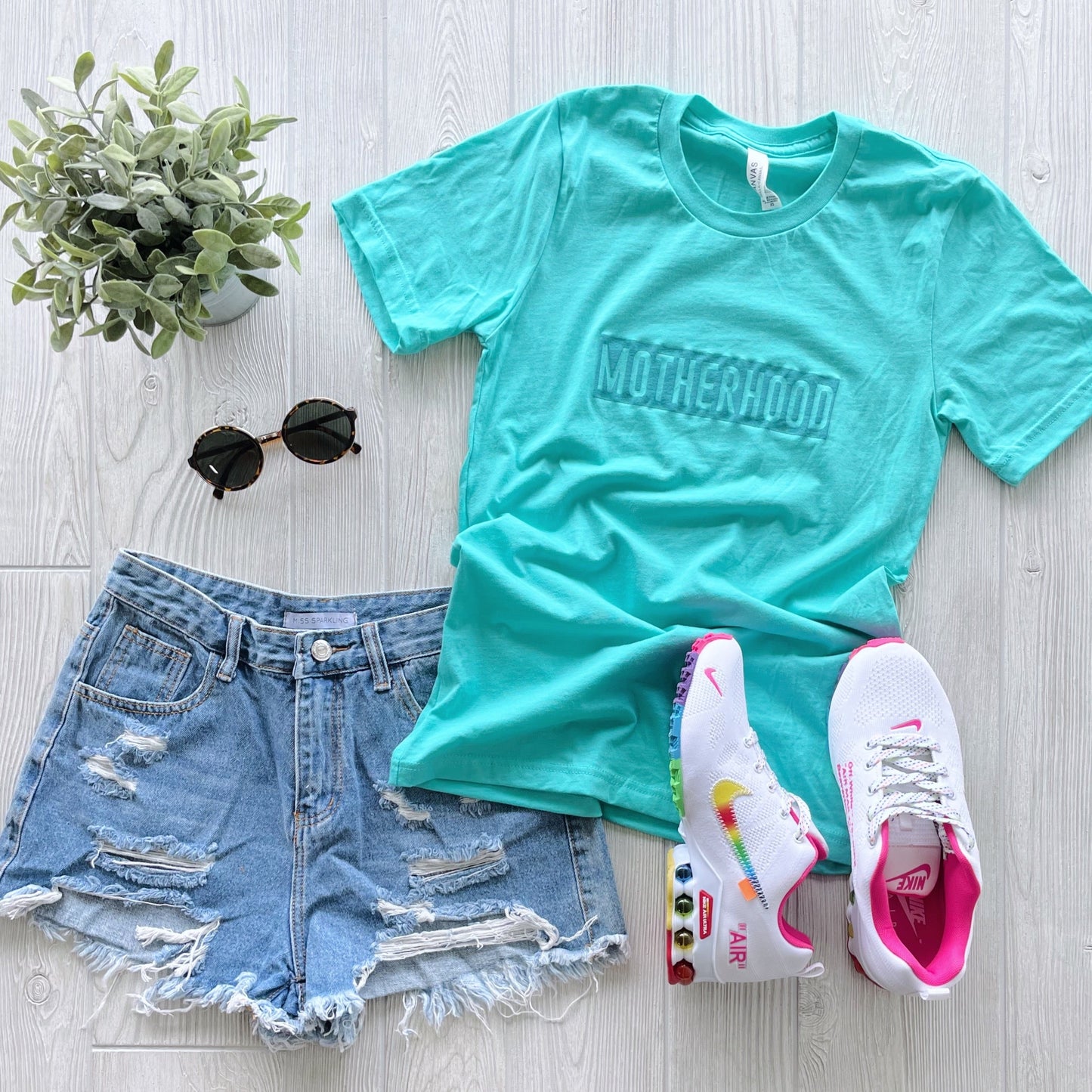 A flat lay of the Motherhood Box • Teal Tonal Tee in unisex fit, paired with distressed denim shorts, colorful-accented white Nike Air sneakers, black sunglasses, and a small potted plant on a light wooden surface.