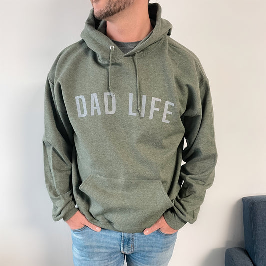 A man wearing the DAD LIFE • Military Green Hoodie stands with his hands in his pockets, looking to the side. He pairs it with light blue jeans.