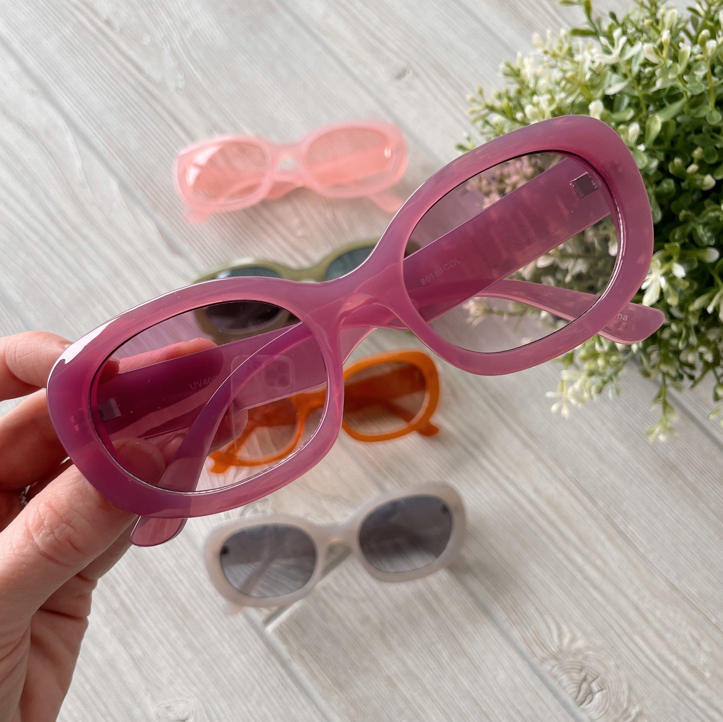 A hand holds pink oval Poppy Sunglasses above a light wooden surface. Three more adult-size pairs—orange, gray, and pink—are below, alongside a green leafy plant on the right. Final sale item. Poppy Sunglasses • Adult • More Colors!.