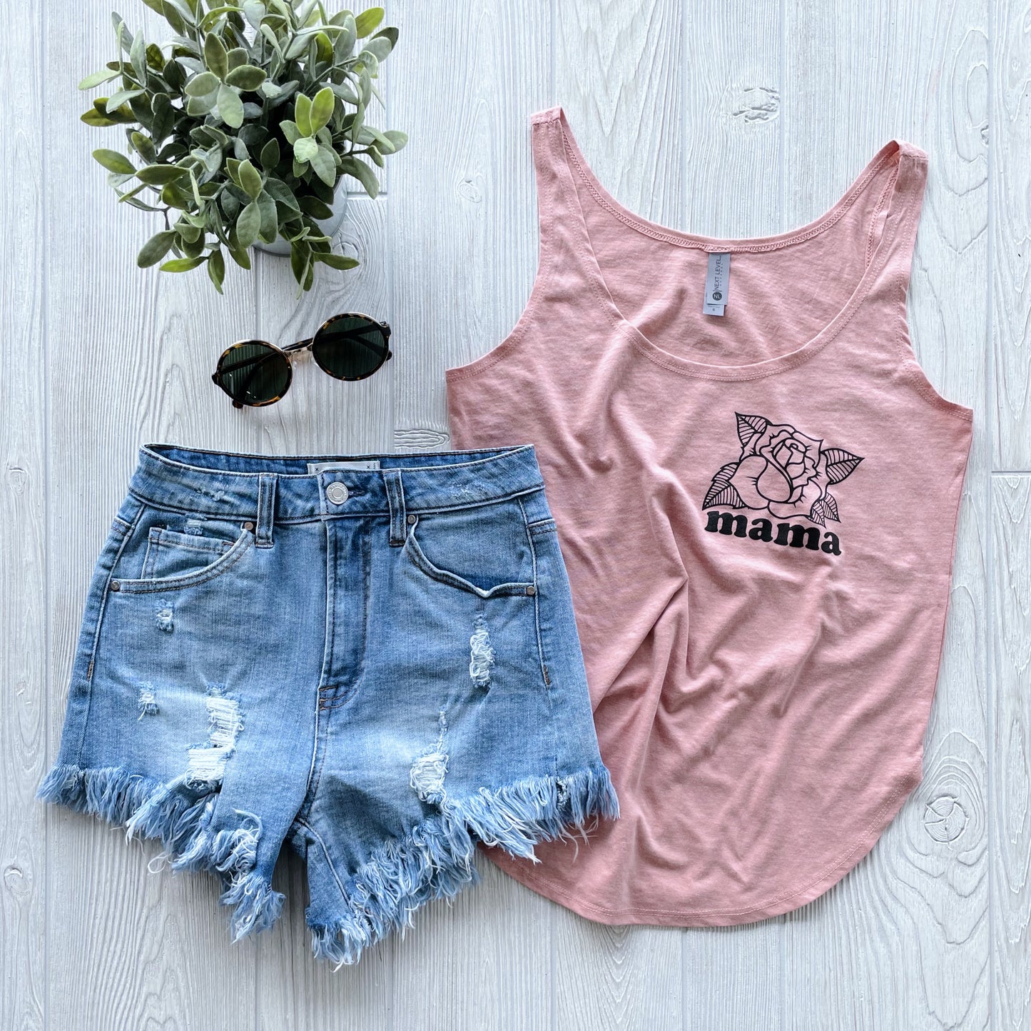 Flat lay of the Mama Rose • Blush Tank, paired with distressed denim shorts, black sunglasses, and a small potted plant on a light wooden surface—a chic choice for women’s style tops.
