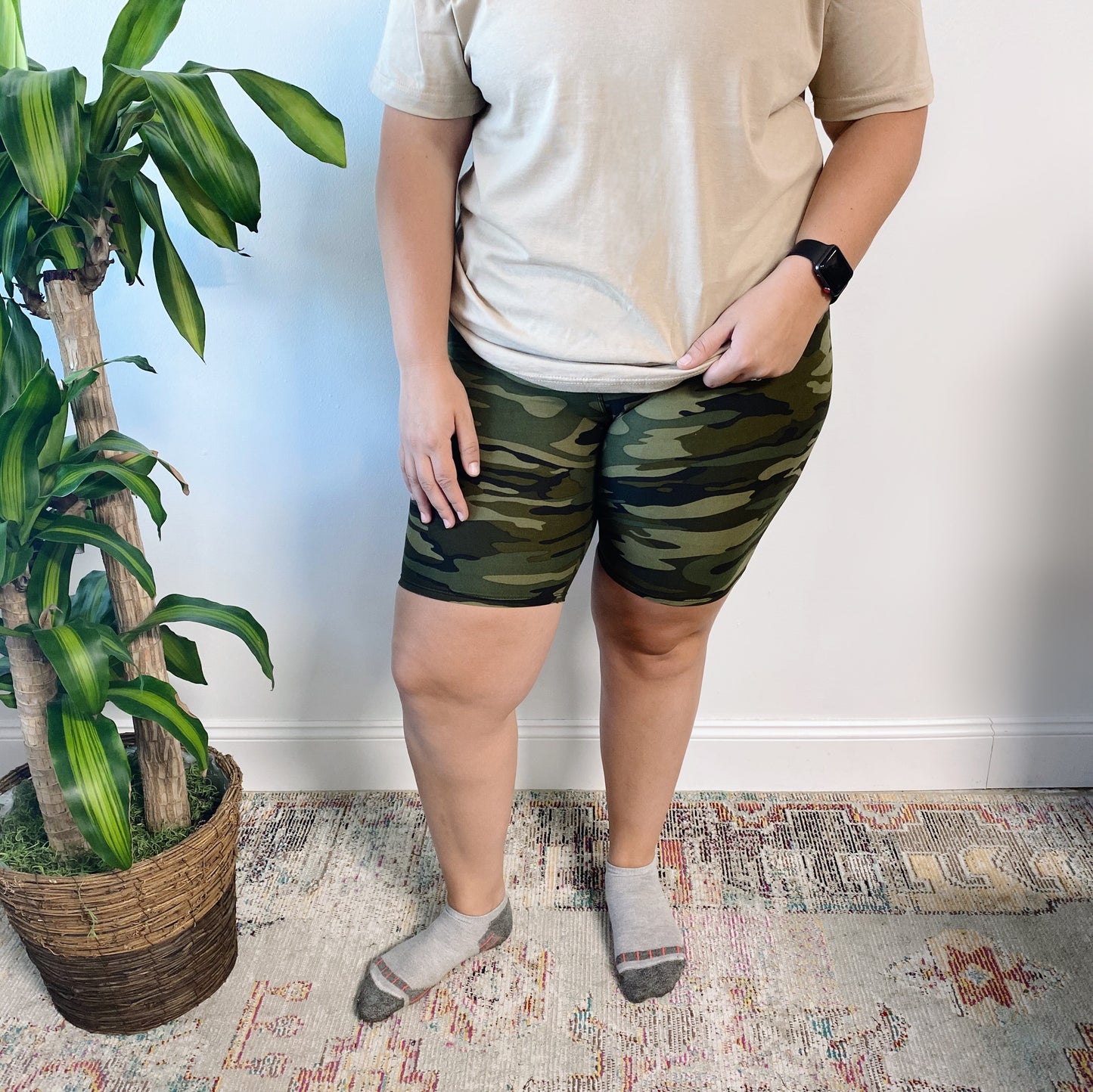 A person wearing a beige shirt, Green Camo • Biker Shorts, and gray socks stands indoors on a patterned rug beside a potted plant and a white wall, with their face not visible.