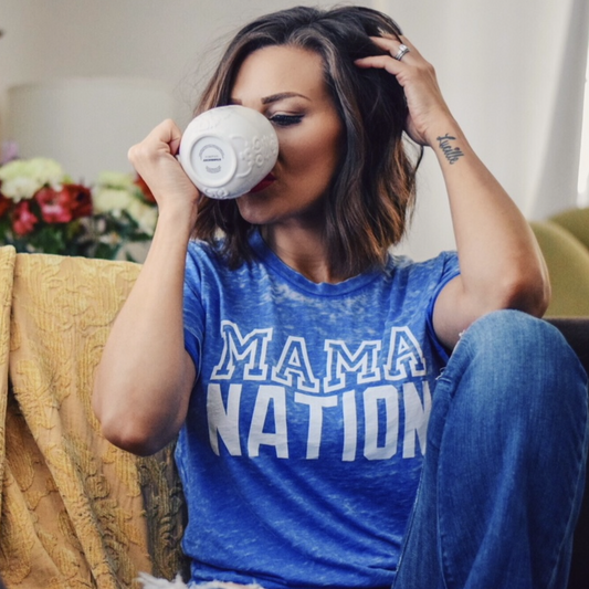 A woman with shoulder-length brown hair wears the Mama Nation • Royal Acid Tee as she sits on a couch, sipping from a white mug and holding her hair back. Flowers and a yellow blanket appear in the background.
