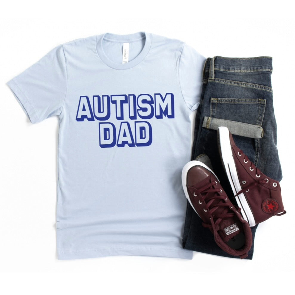 The Autism Dad • Adult Tee, a light blue shirt with bold blue AUTISM DAD print, inspired by a World Autism Month fundraiser, is styled with dark jeans and maroon sneakers on a white background.