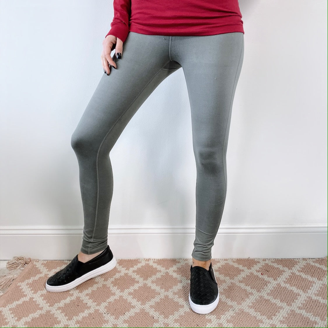 A person wearing a red long-sleeve shirt, Basic Sage Leggings • 6168, and black slip-on shoes stands on a beige patterned rug against a white wall. Their face is not visible.
