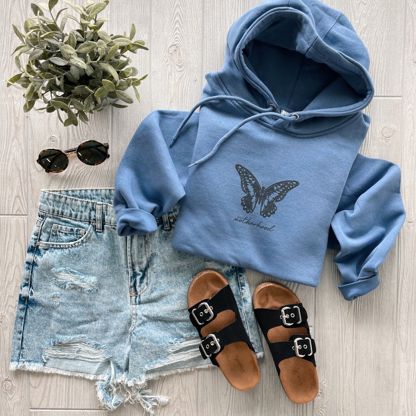 Flat lay of the Butterfly Motherhood • Periwinkle Hoodie with fleece lining, paired with distressed denim shorts, black buckle sandals, round sunglasses, and a small potted plant on a light wooden floor.