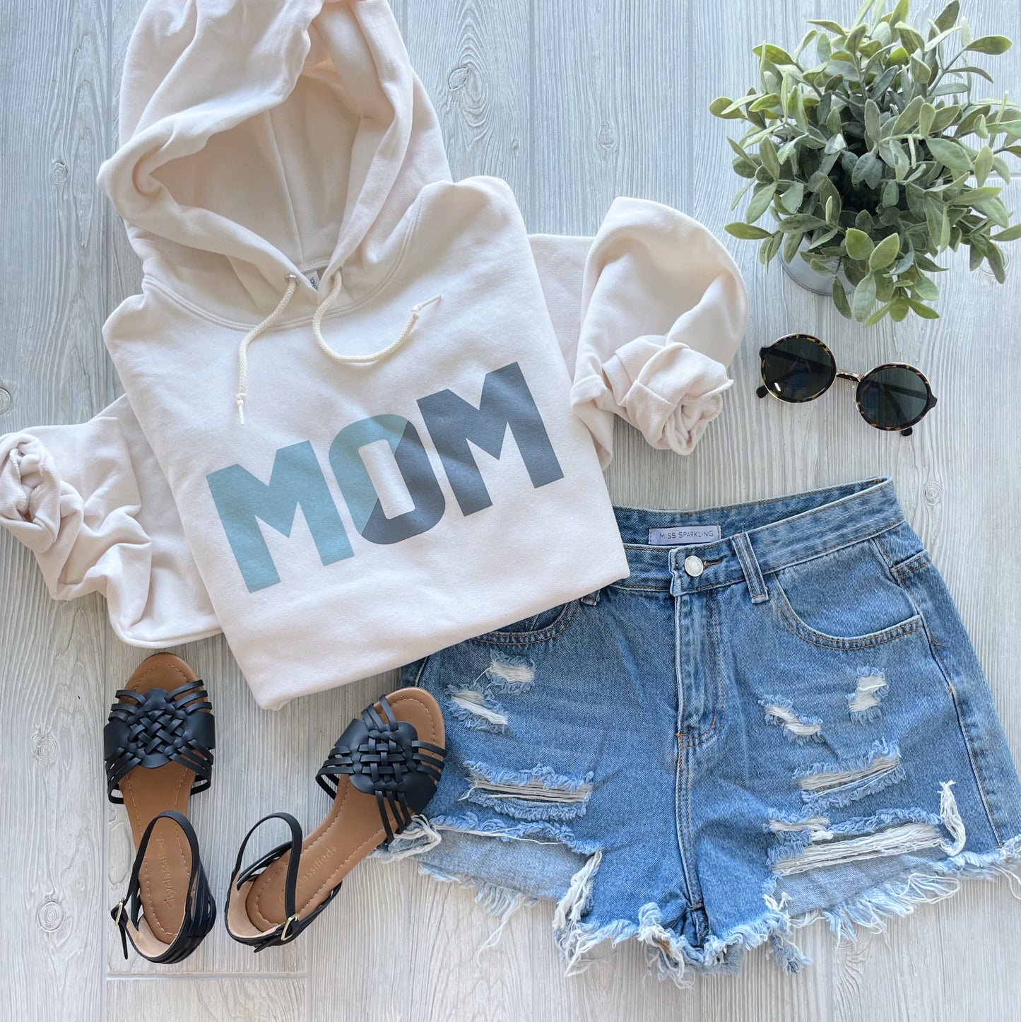 A flat lay of the MOM Slice Print • Sweet Cream Hoodie, distressed denim shorts, black sandals, round sunglasses, and a small green potted plant on a light wooden surface.
