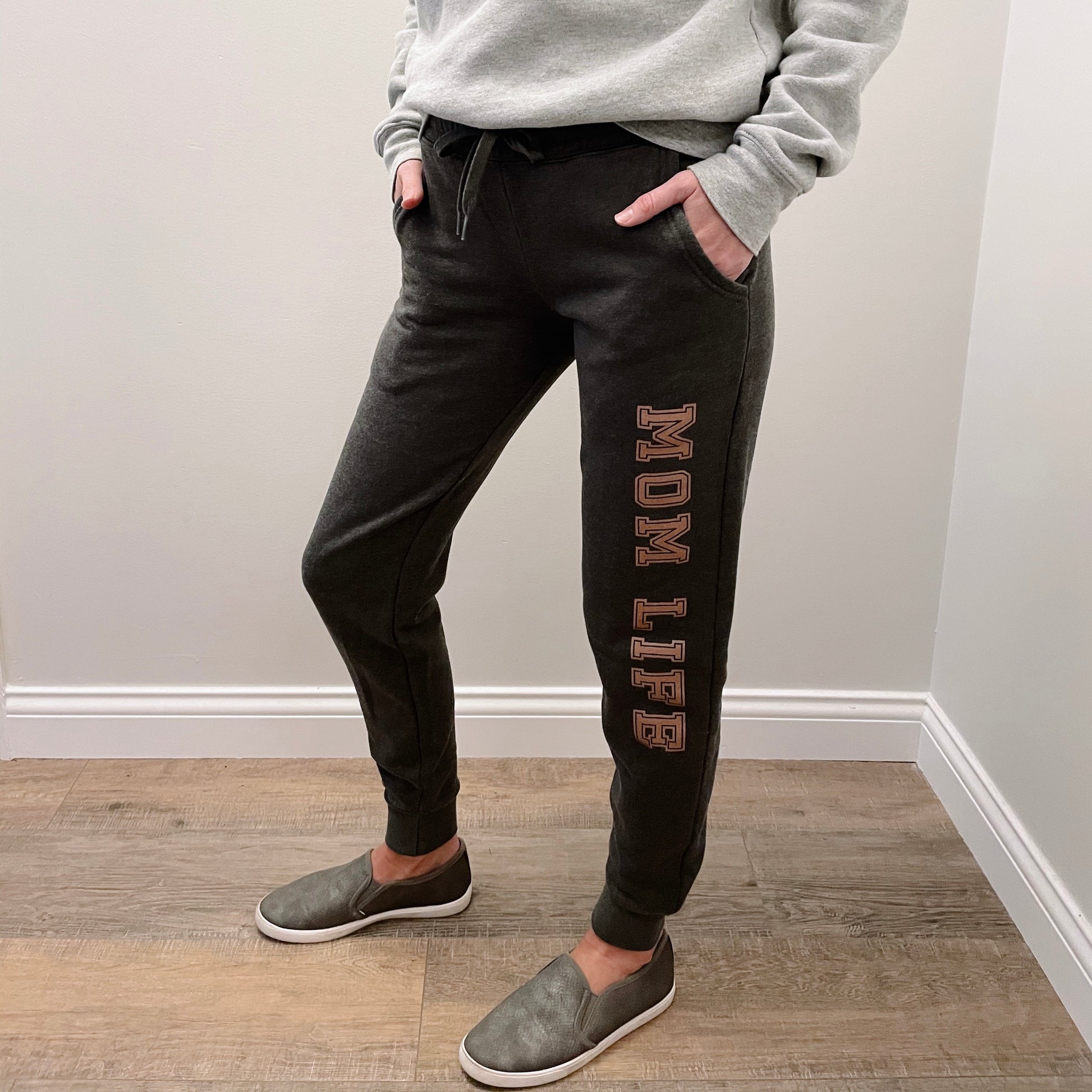 Wearing MOM LIFE • Charcoal Jogger pants, a light gray sweatshirt, and slip-on shoes, a person stands with hands in pockets on a wood floor against a white wall.