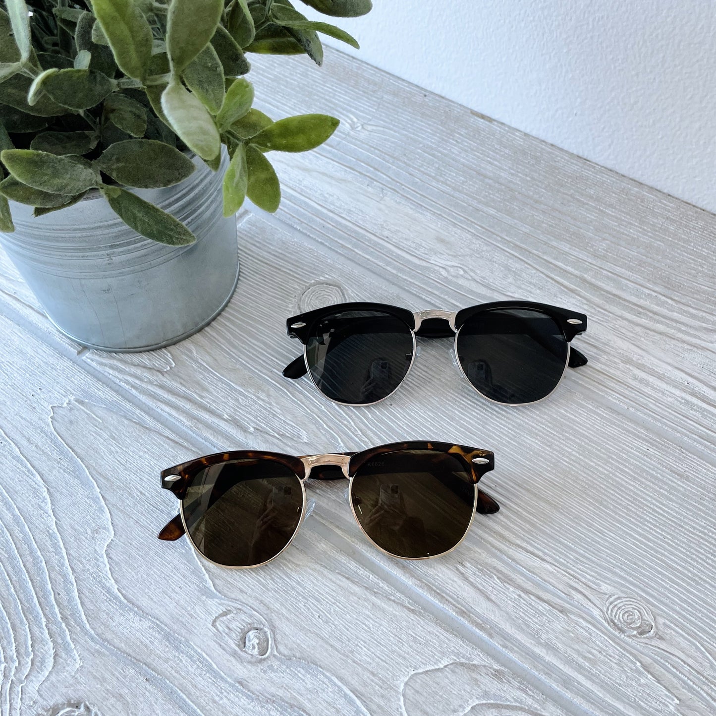 Two pairs of Parker Sunnies • Kids, one in black and one in tortoiseshell, rest on a light wooden table beside a potted green plant in a metal container. Both pairs offer UV protection for stylish safety.