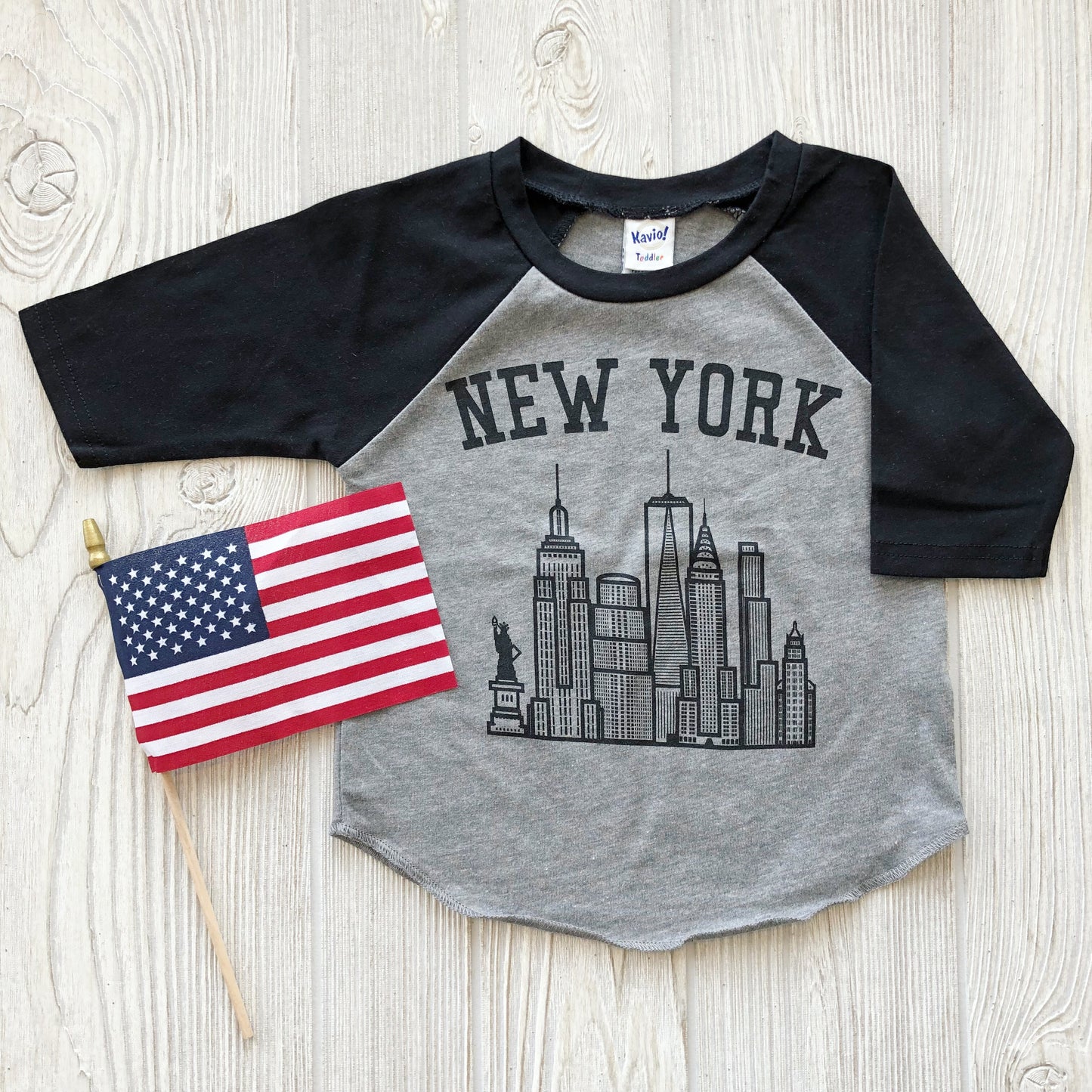 The New York • Raglan, a gray and black top featuring a bold city skyline design, is shown on a wooden surface next to a small American flag on a stick.