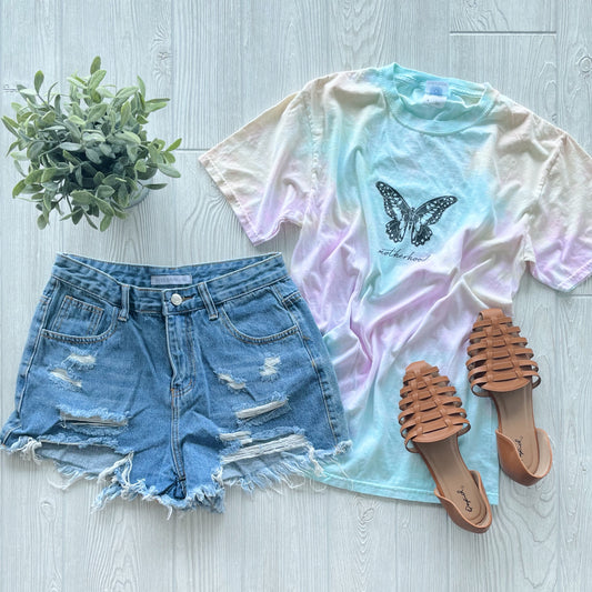 A Butterfly • Pastel Tie Dye Tee pairs with distressed denim shorts, tan woven sandals, and a small potted plant, all styled on a light wood floor.