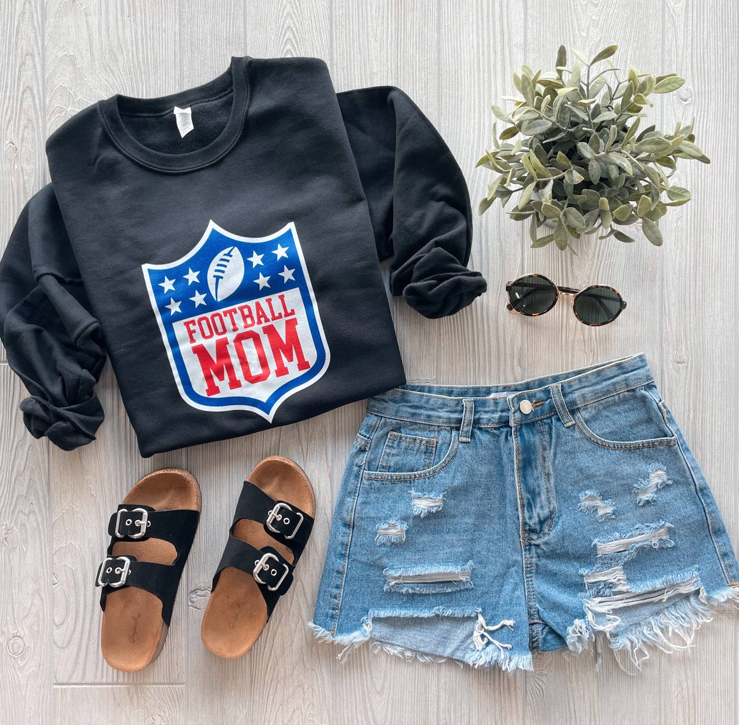 The Football Mom • Black Pullover from the Sports Mom Collection features a football logo and is styled with distressed shorts, black sandals, round sunglasses, and a potted plant on a light wood surface.