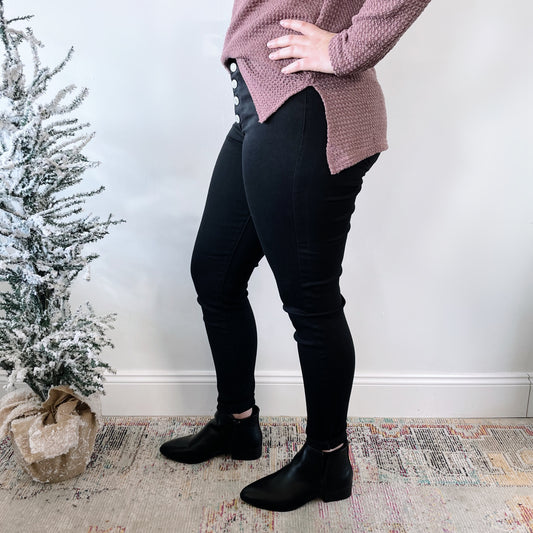 Indoors, a person wears mauve textured sweater, Phalesha Jeggings SIZE 0 & 1—true to fit high-waisted black pants with buttons—and black ankle boots. Their hand is on their hip near a frosted artificial tree on a patterned rug.