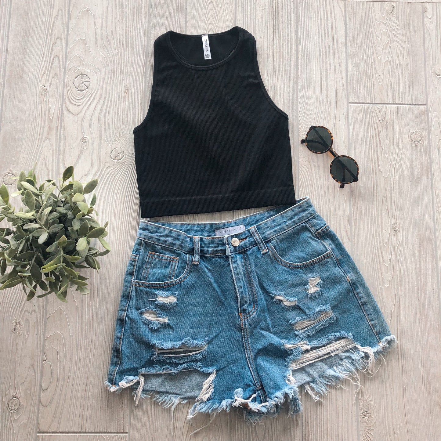 The Sophia High-Neck Crop in black is styled with distressed denim shorts, round black sunglasses, and a small green faux plant, all arranged on a light wood floor.