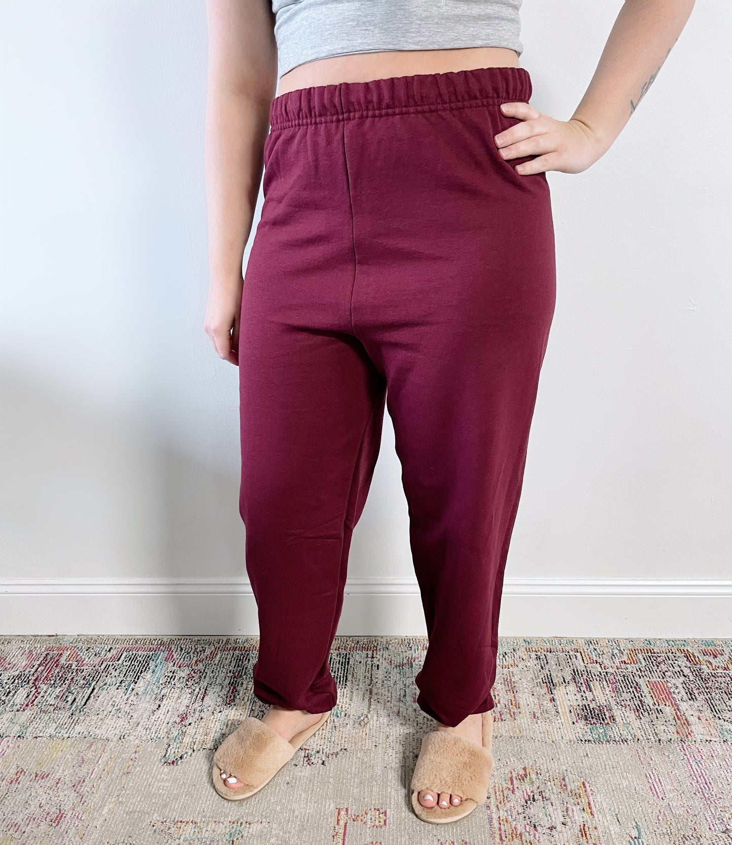 A person in a gray crop top, Mamacita Sweats • Maroon, and tan fuzzy slippers stands on a patterned rug by a white wall. Their left hand rests on their hip, highlighting cozy loungewear style.