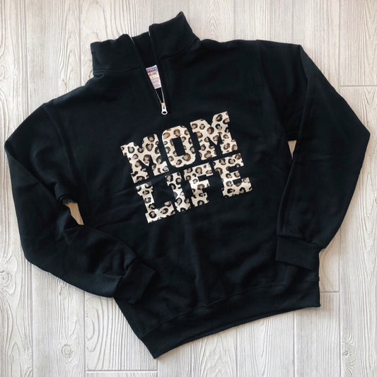 The Mom Life Cheetah • Quarter Zip is a black pullover sweatshirt featuring bold cheetah print MOM LIFE lettering on the front, shown laid flat on a light wooden floor.