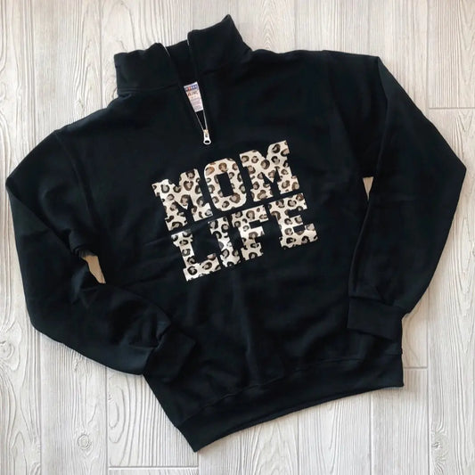 The Mom Life Cheetah • Quarter Zip is a black pullover sweatshirt featuring bold cheetah print MOM LIFE lettering on the front, shown laid flat on a light wooden floor.