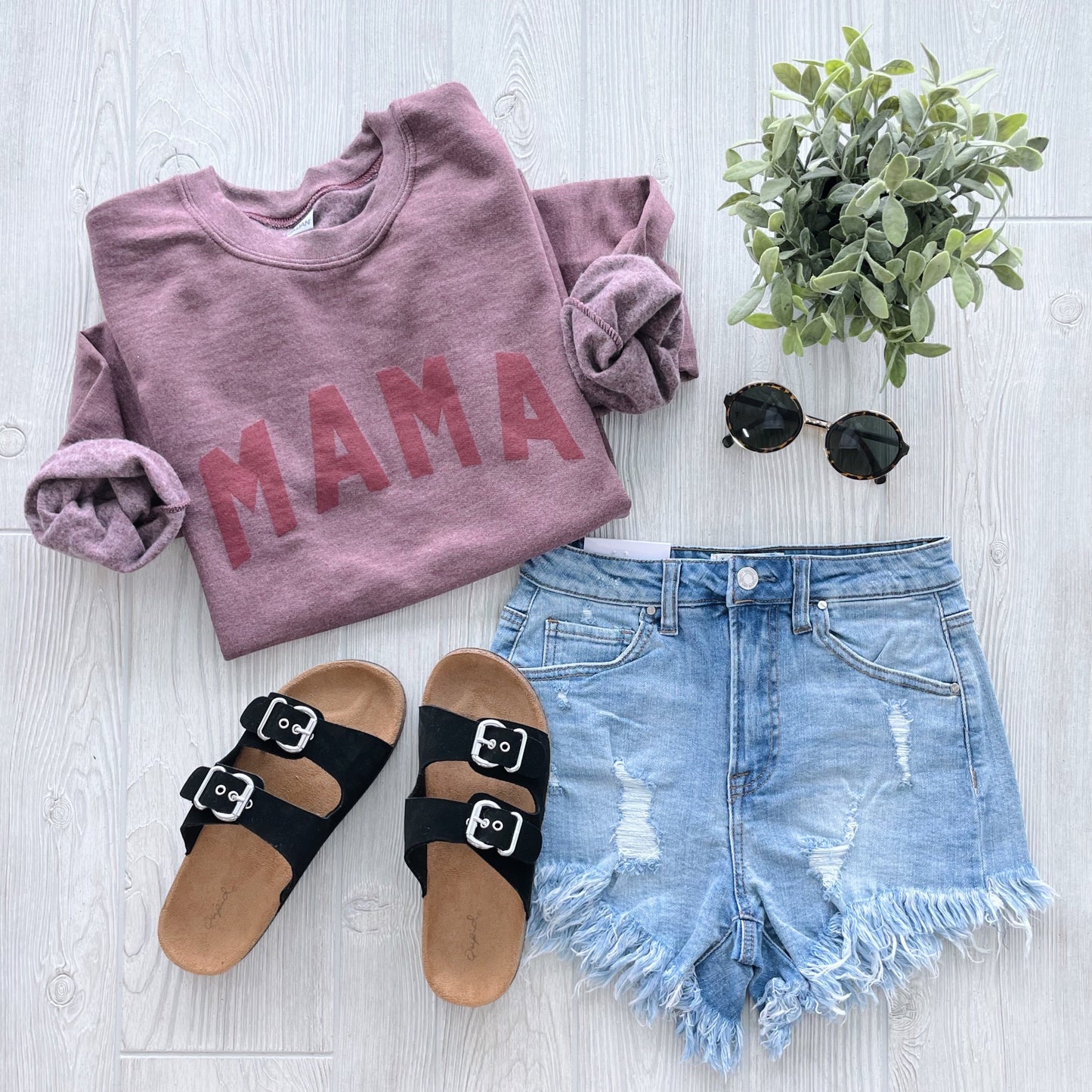 A flat lay of the MAMA • Tonal Maroon Pullover with distressed denim shorts, black buckle sandals, round sunglasses, and a small potted plant on a light wooden surface—perfect for cozy unisex style fans.