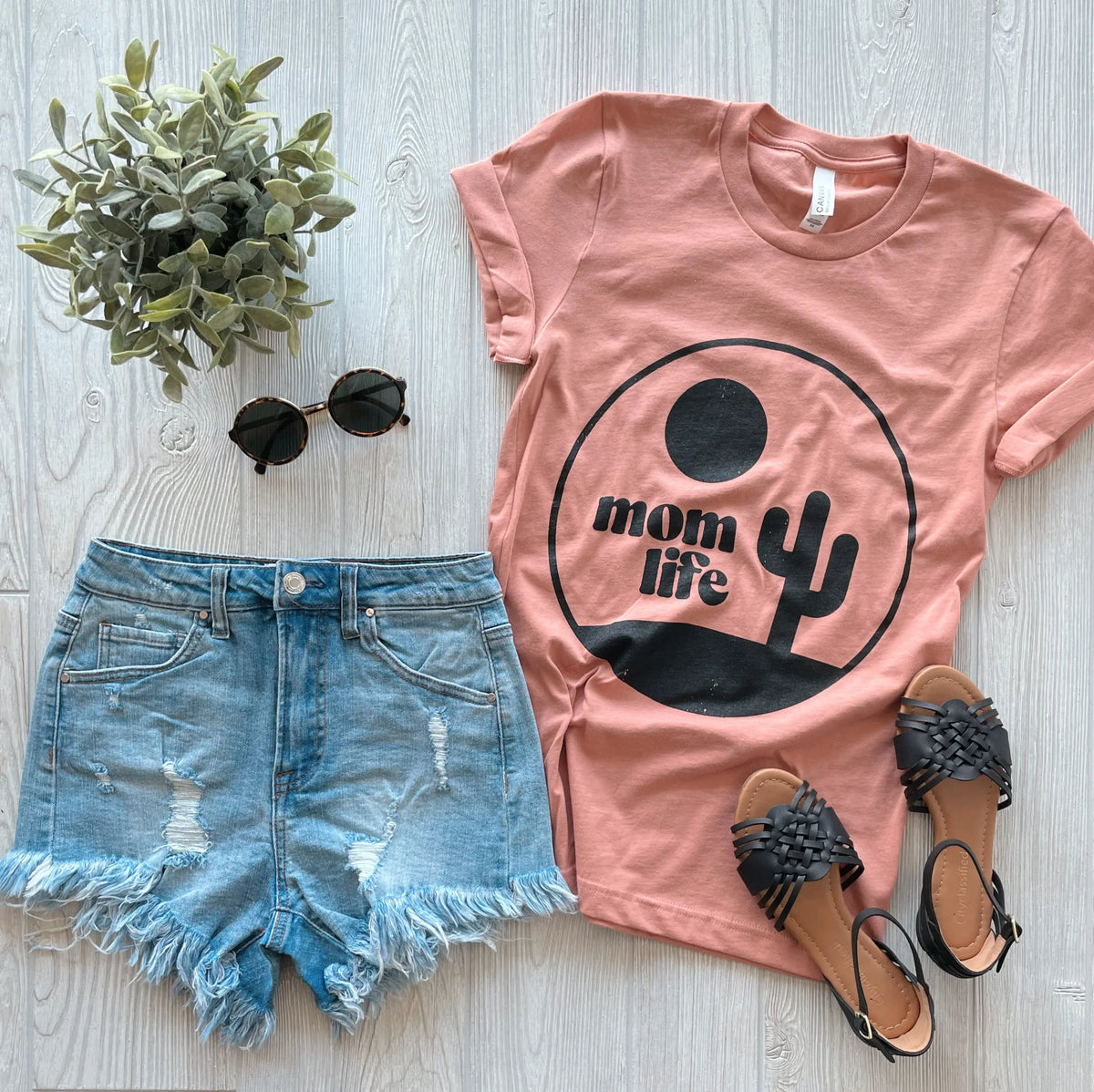 Flat lay of the Cactus Mom Life • Terracotta Tee, styled with distressed denim shorts, black woven sandals, round sunglasses, and a small potted plant—an effortless unisex fit on a light wood background.