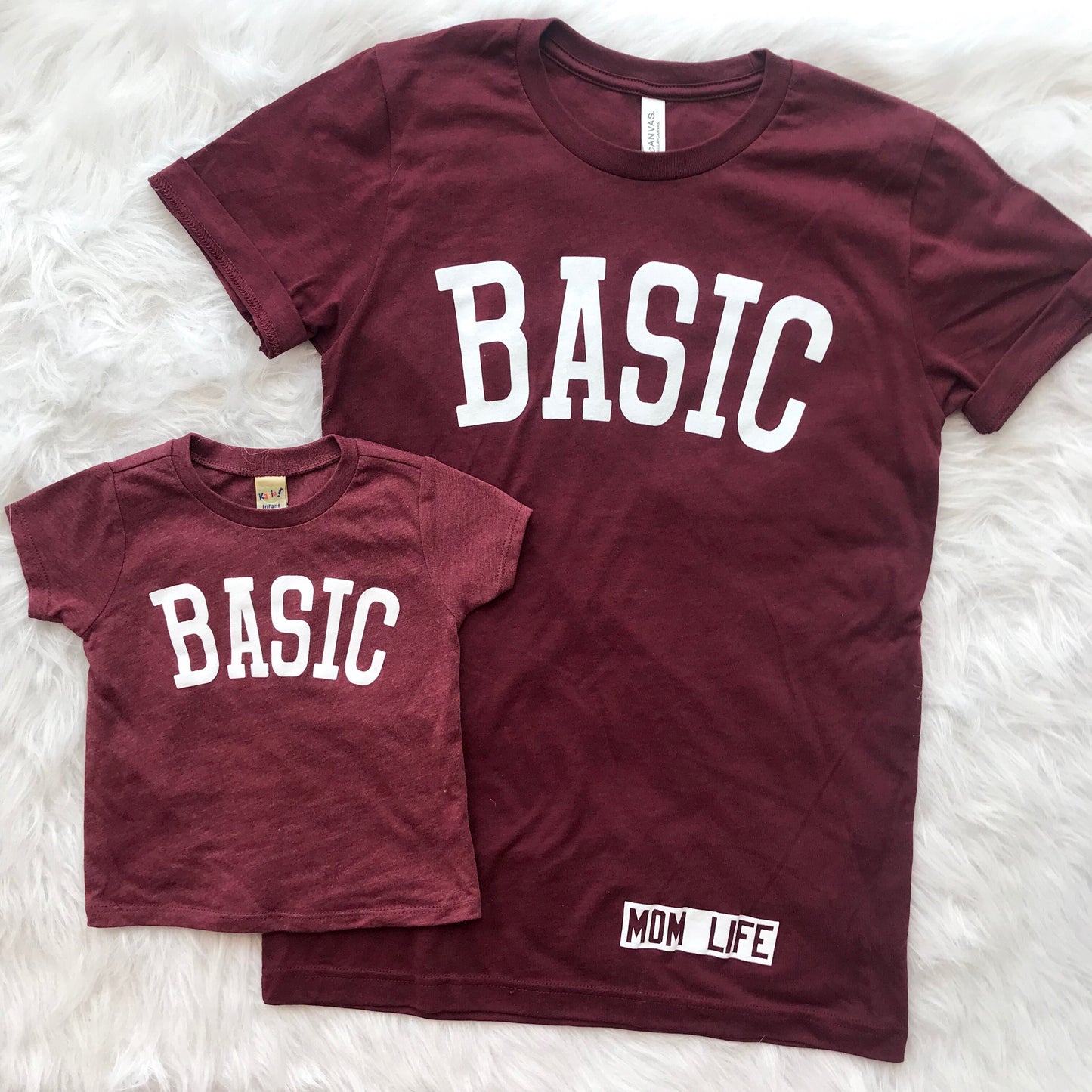 Two maroon BASIC Tee • Maroon shirts are shown on a white fluffy background. The larger unisex shirt has BASIC in bold white letters with a MOM LIFE patch, and the smaller tee also features BASIC in white.