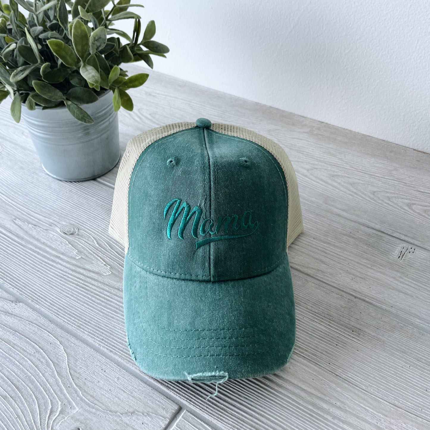 The Distressed Mama Hat • Pine, a green and beige canvas cap with Mama embroidered on the front, sits on a light wooden surface beside a small potted plant with green leaves.