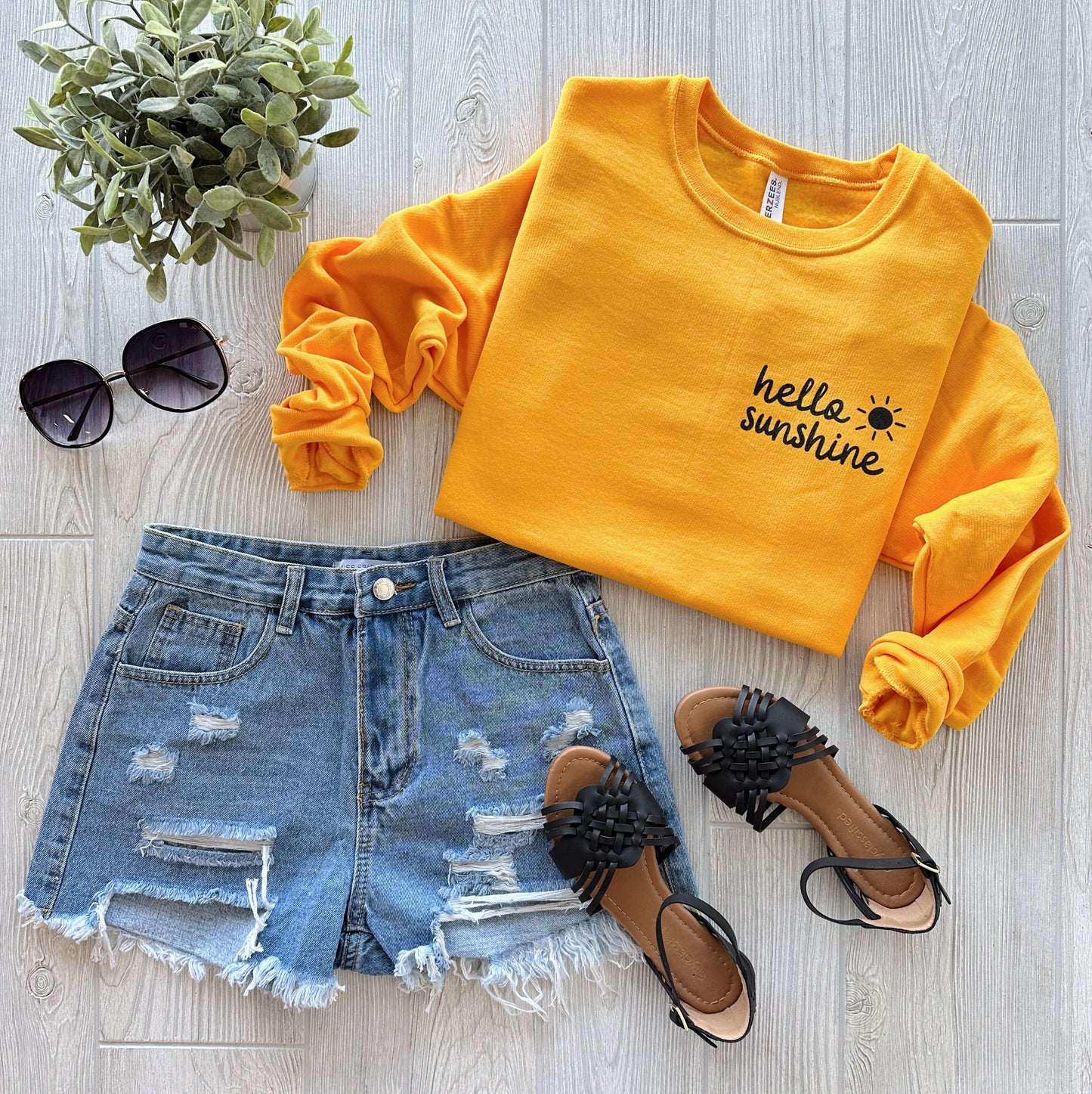 Flat lay of the Hello Sunshine • Golden Pullover in a comfy fit, paired with distressed denim shorts, black sandals, sunglasses, and a small green plant on a light wood background.