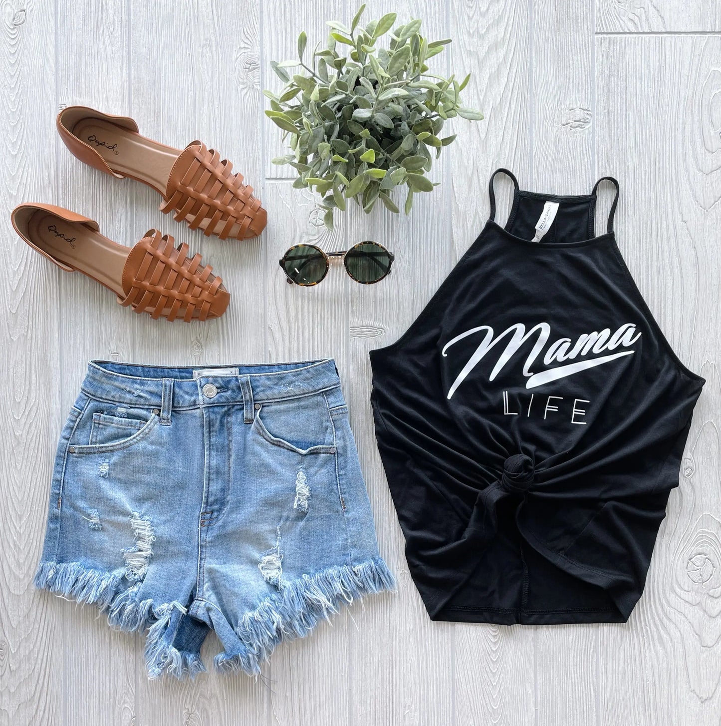 Flat lay of a casual summer outfit: brown woven slip-on shoes, round sunglasses, the Retro Mama • High Neck Tank with vintage Mama Life text, distressed denim shorts with frayed hems, and a small potted plant.