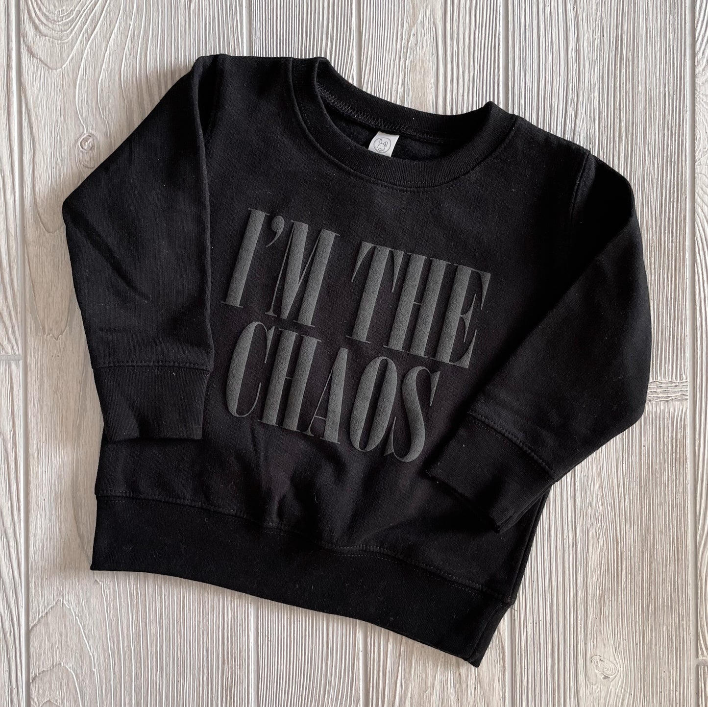 The CHAOS • Kids Pullover is a black sweatshirt with IM THE CHAOS in bold letters, shown on a light wood surface. Perfect for adding attitude to your kiddos closet, plus a matching adult version for coordinated style.