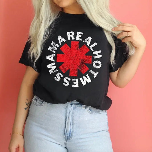 A woman with long blonde hair wears the REAL HOT MESS MAMA black tee, featuring a bold red asterisk and “MAMA REAL HOT MESS” in white, styled with light blue jeans against a pink background.