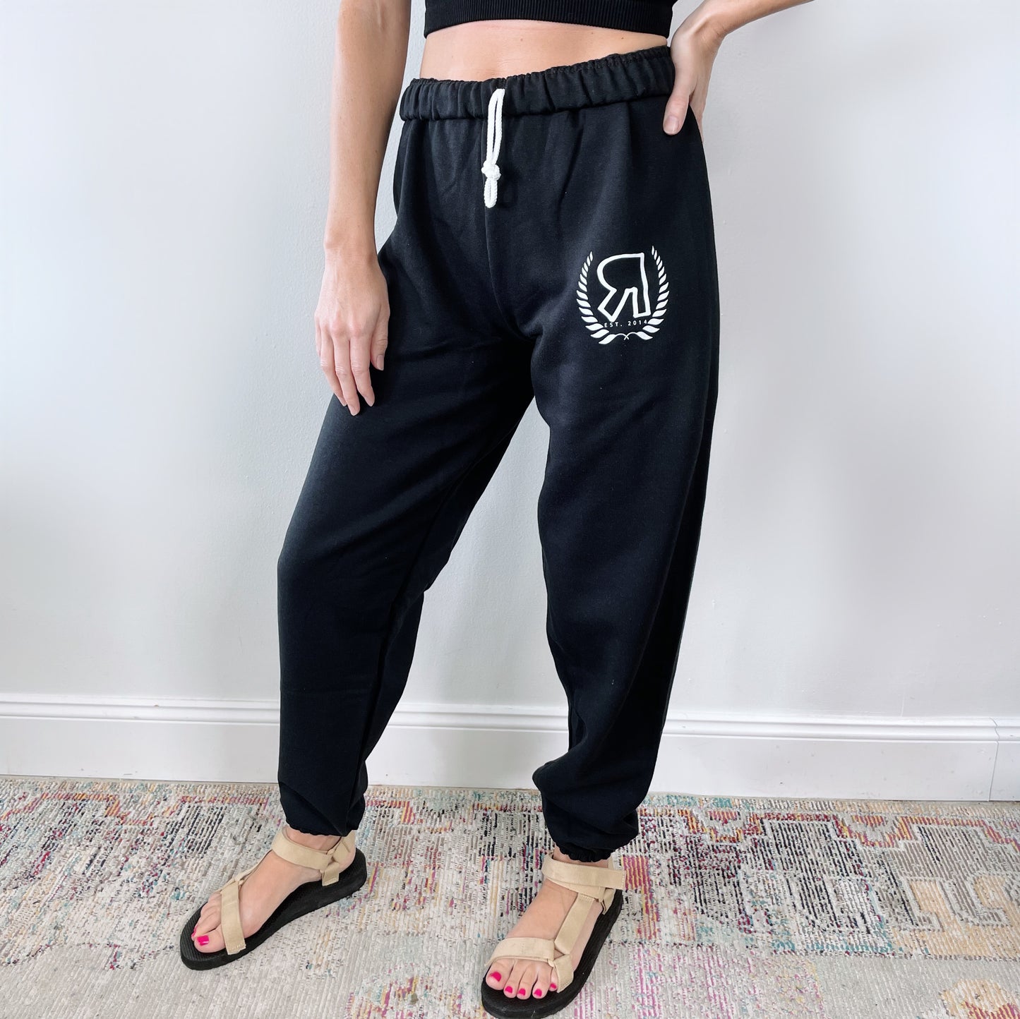 A person stands indoors on a colorful rug, showing only their lower half, wearing black Boyfriend Sweats • Logo with a white logo on the left thigh, a black crop top, and beige sandals in front of a white wall.