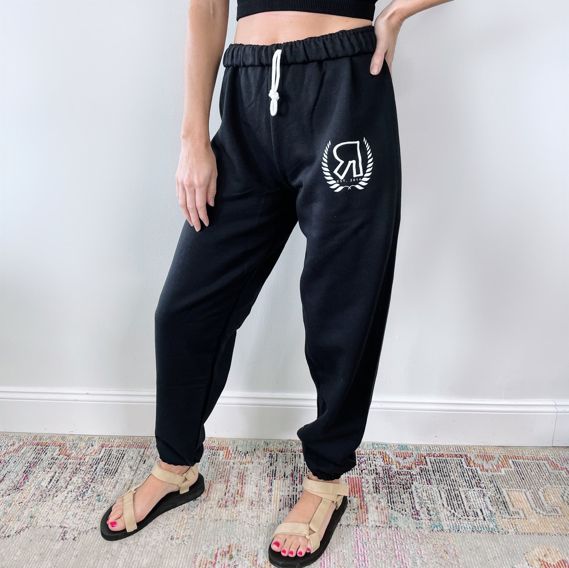 A person stands indoors on a colorful rug, showing only their lower half, wearing black Boyfriend Sweats • Logo with a white logo on the left thigh, a black crop top, and beige sandals in front of a white wall.