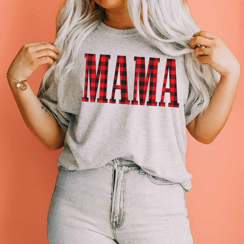 A woman with long blonde hair wears the MAMA • Buffalo Plaid tee, featuring a bold red and black plaid MAMA print, standing against a peach-colored background.