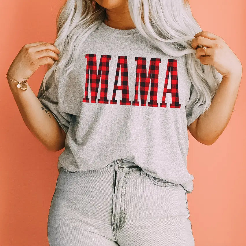 A woman with long blonde hair wears the MAMA • Buffalo Plaid tee, featuring a bold red and black plaid MAMA print, standing against a peach-colored background.