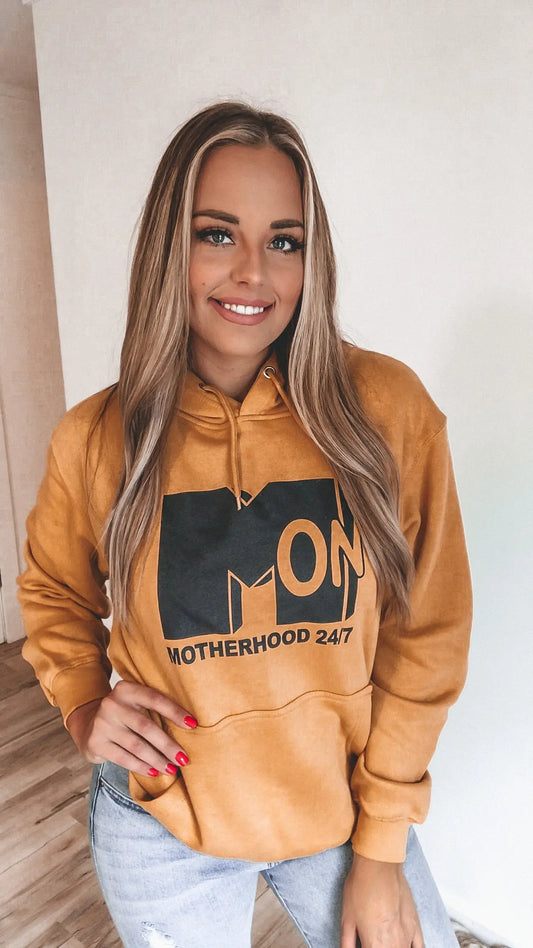 A smiling woman with long blonde hair models the Motherhood 24/7 Mustard Hoodie, featuring bold black lettering and a cozy fleece lining—an ideal choice for stylish moms, shown indoors against a white wall and wooden floor.