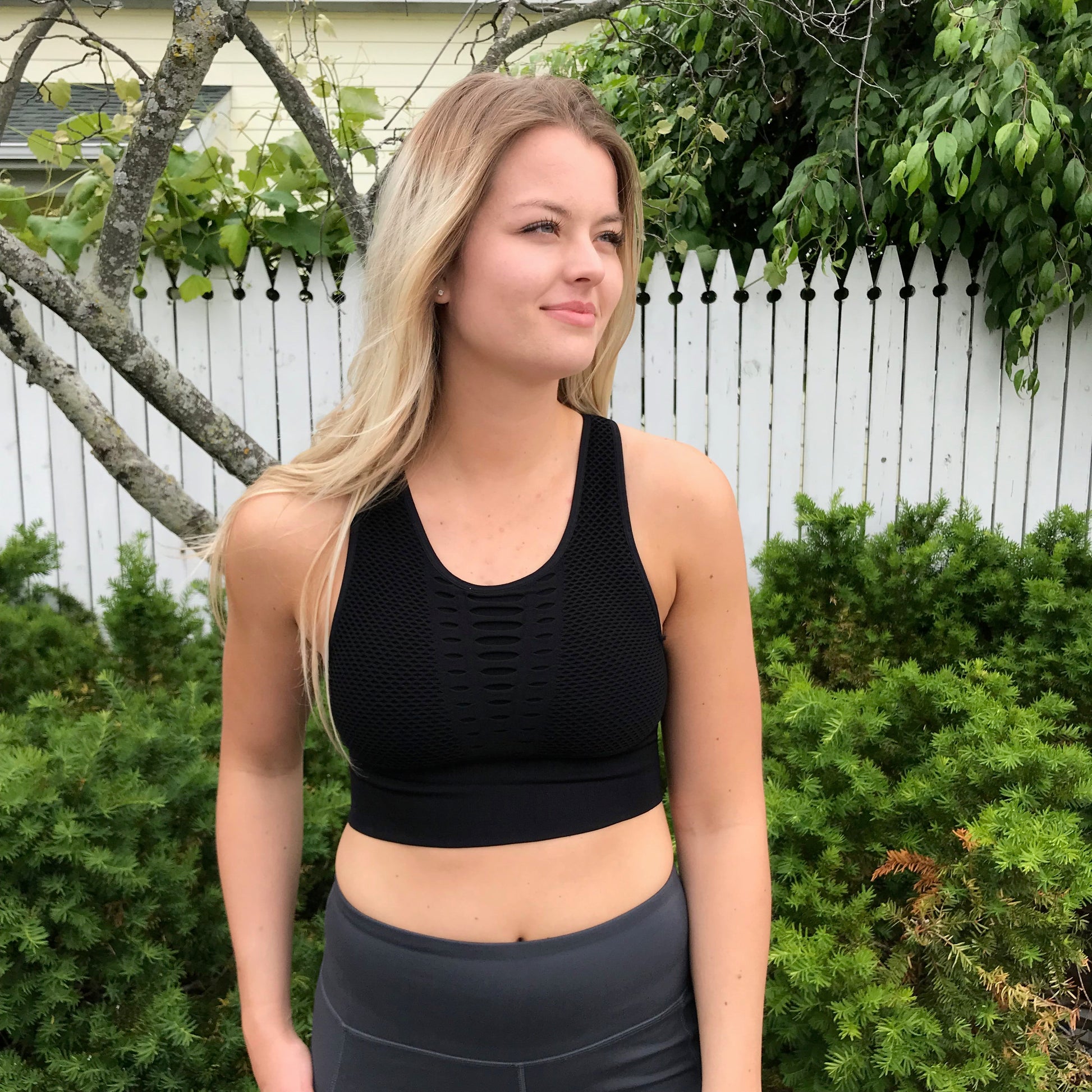 A woman with long blonde hair wears the Grated Sports Bra in black, size small—true to size for a flattering fit—while standing outdoors by a white picket fence and green bushes, looking off to the side with a slight smile.