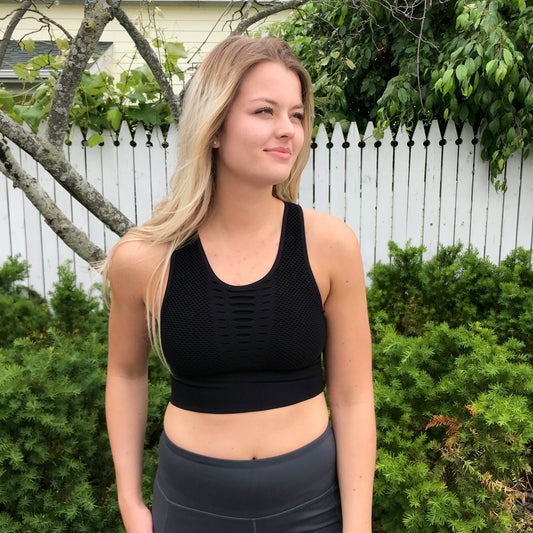 A woman with long blonde hair wears the Grated Sports Bra in black, size small—true to size for a flattering fit—while standing outdoors by a white picket fence and green bushes, looking off to the side with a slight smile.