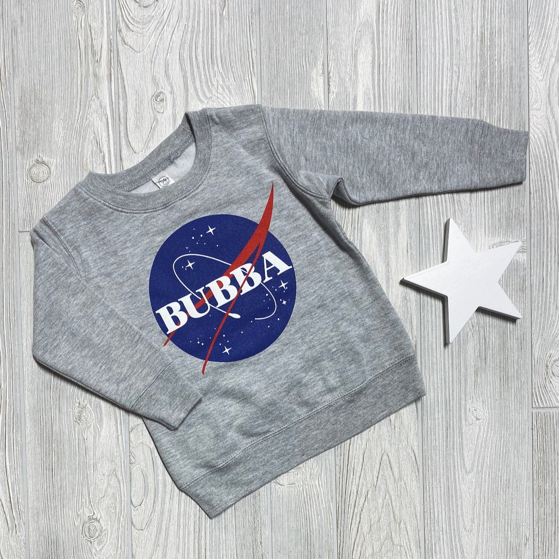 A comfy unisex Bubba Space • Pullover kids sweatshirt in gray, featuring a NASA-inspired logo, is laid on a light wooden floor next to a white star-shaped decoration.