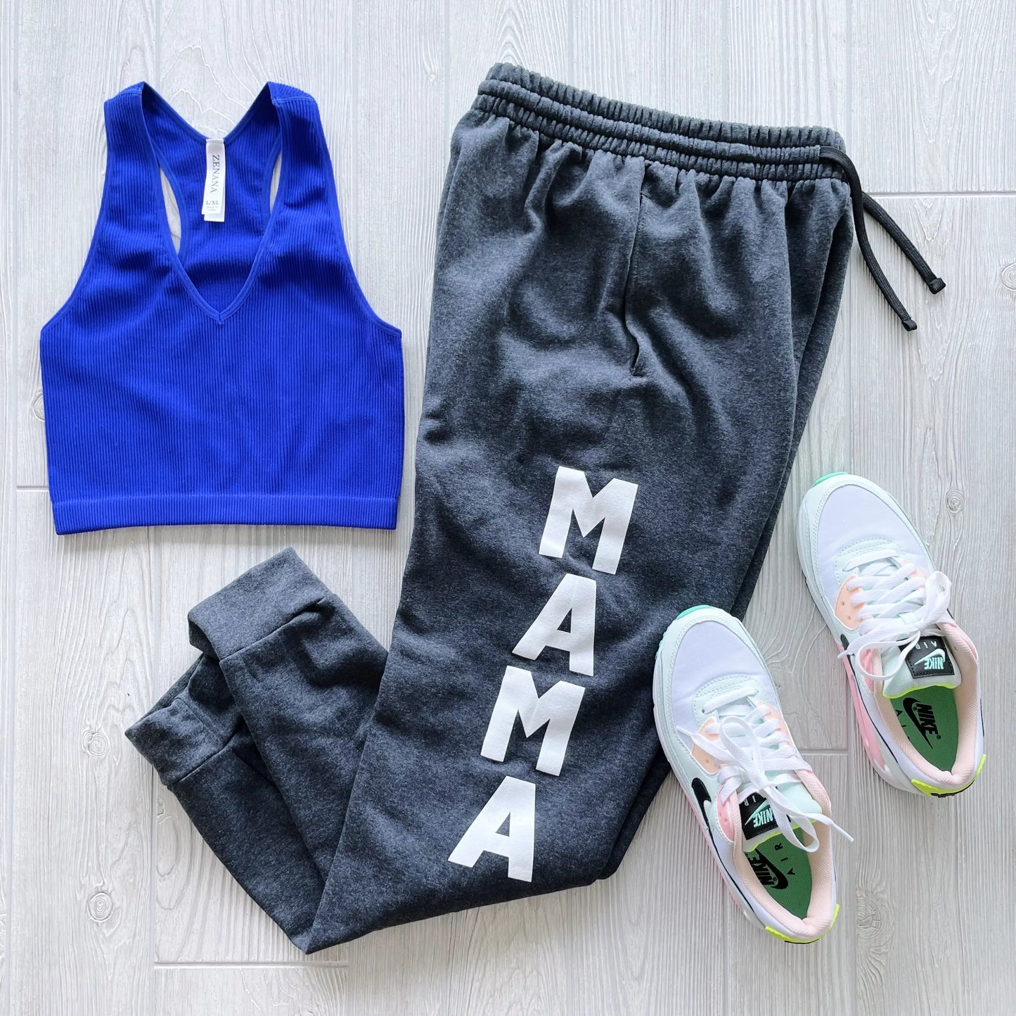 A blue sports bra, New Mama Joggers in Black Heather, and white Nike sneakers with pastel green and pink accents create a cozy loungewear look on a light wood floor.