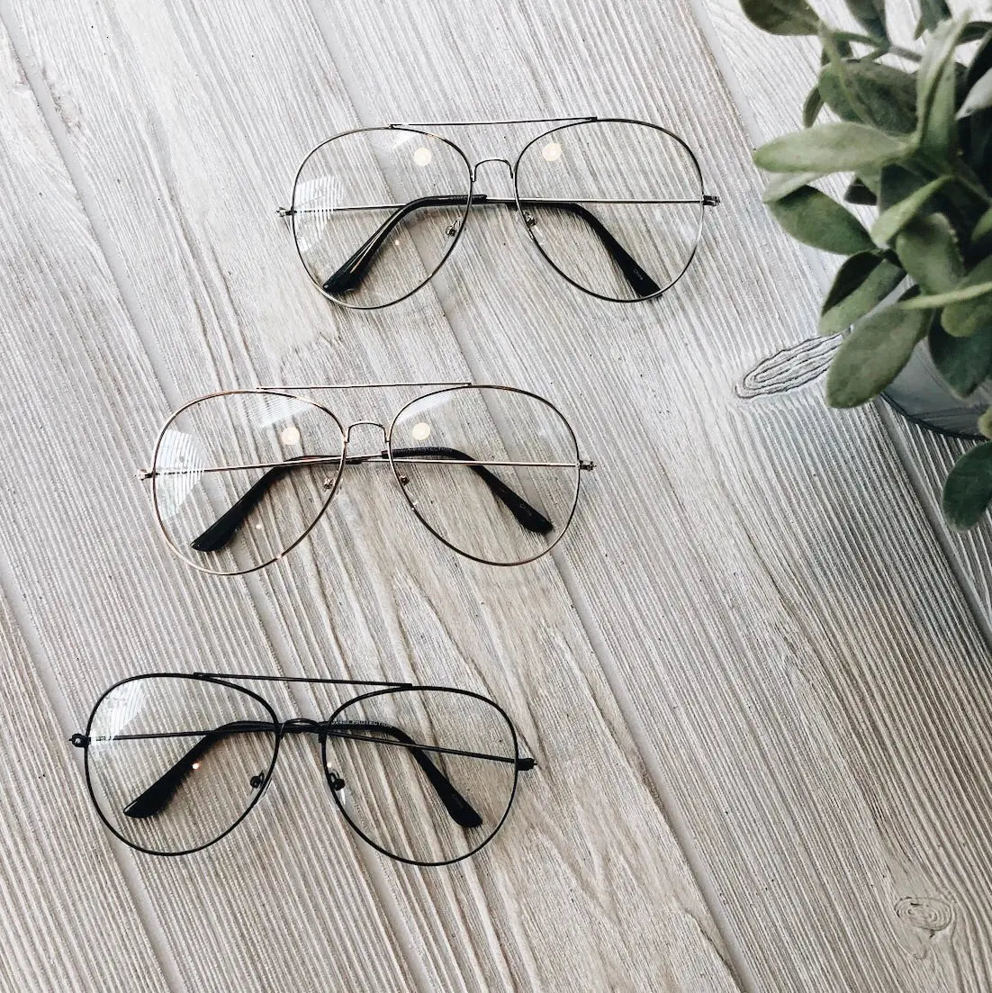 Three pairs of Alex • Blue Light Glasses • More Colors! with metal frames are displayed on a light wooden surface next to a small green potted plant, perfect for reducing eye strain.