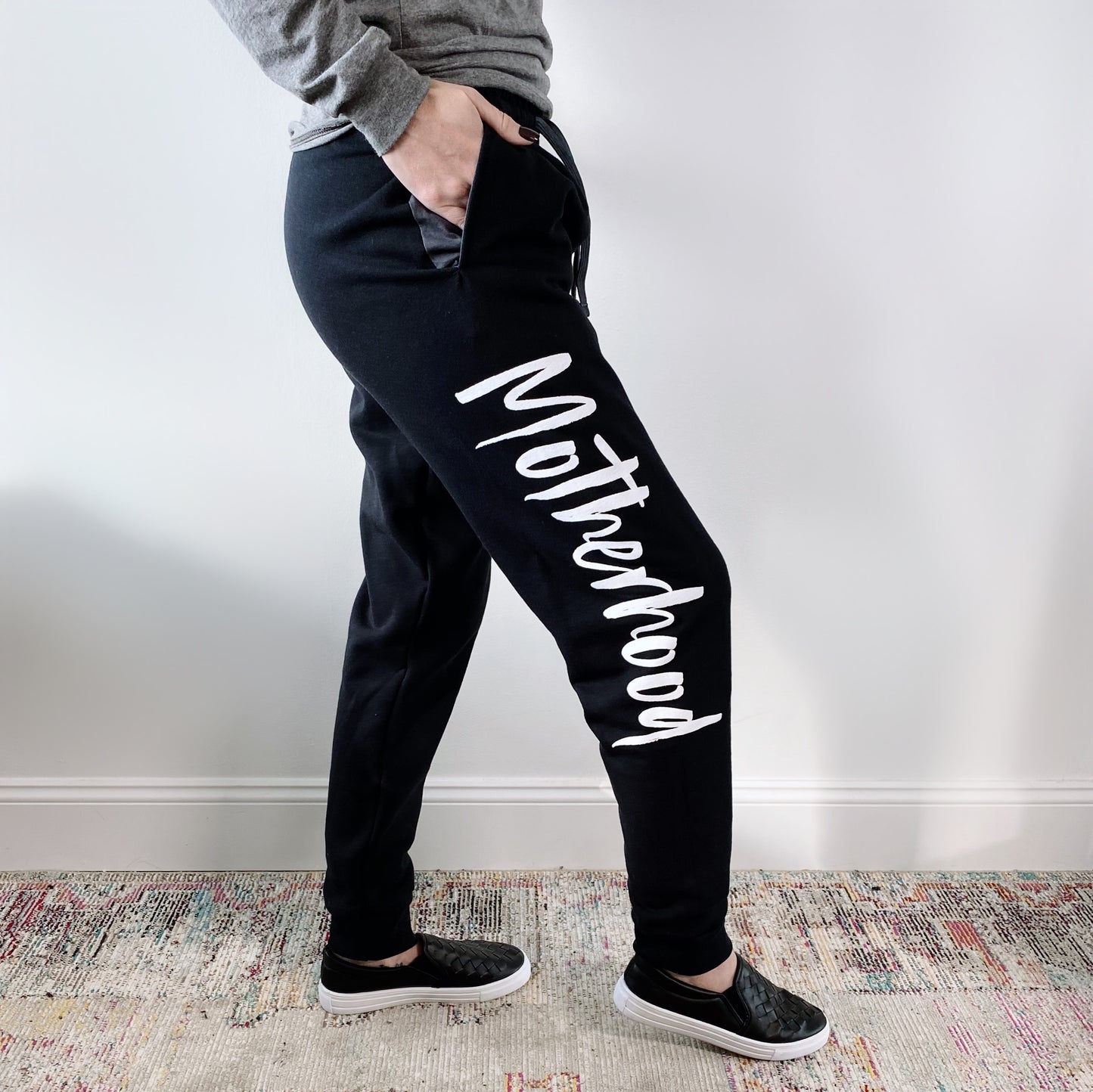A person stands sideways against a white wall wearing the Motherhood Jogger • Black, featuring a bold Motherhood print down the leg, paired with a gray top and black slip-on shoes.