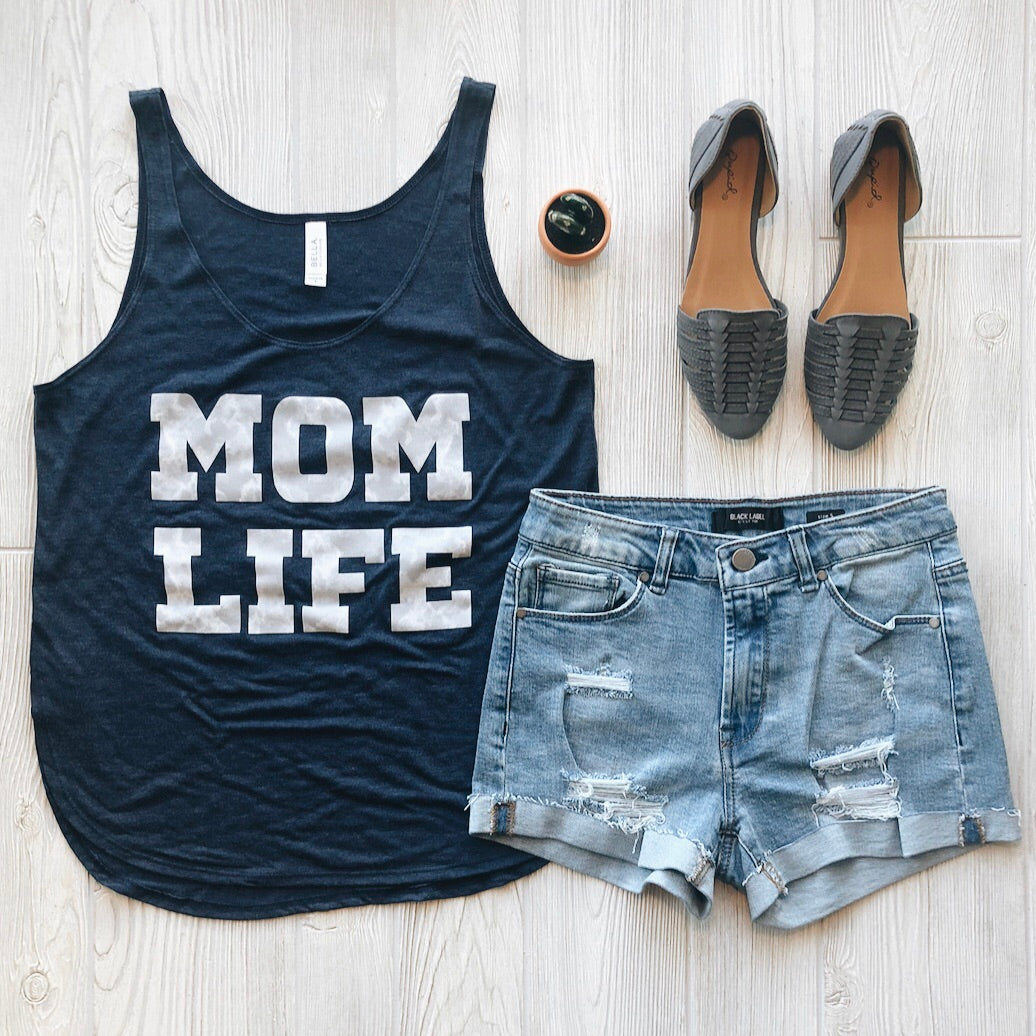 A Smoke Print • Side Slit Tank with a flowy fit is styled with distressed denim shorts, gray woven slip-on shoes, and a small round container, all displayed on a light wood floor.