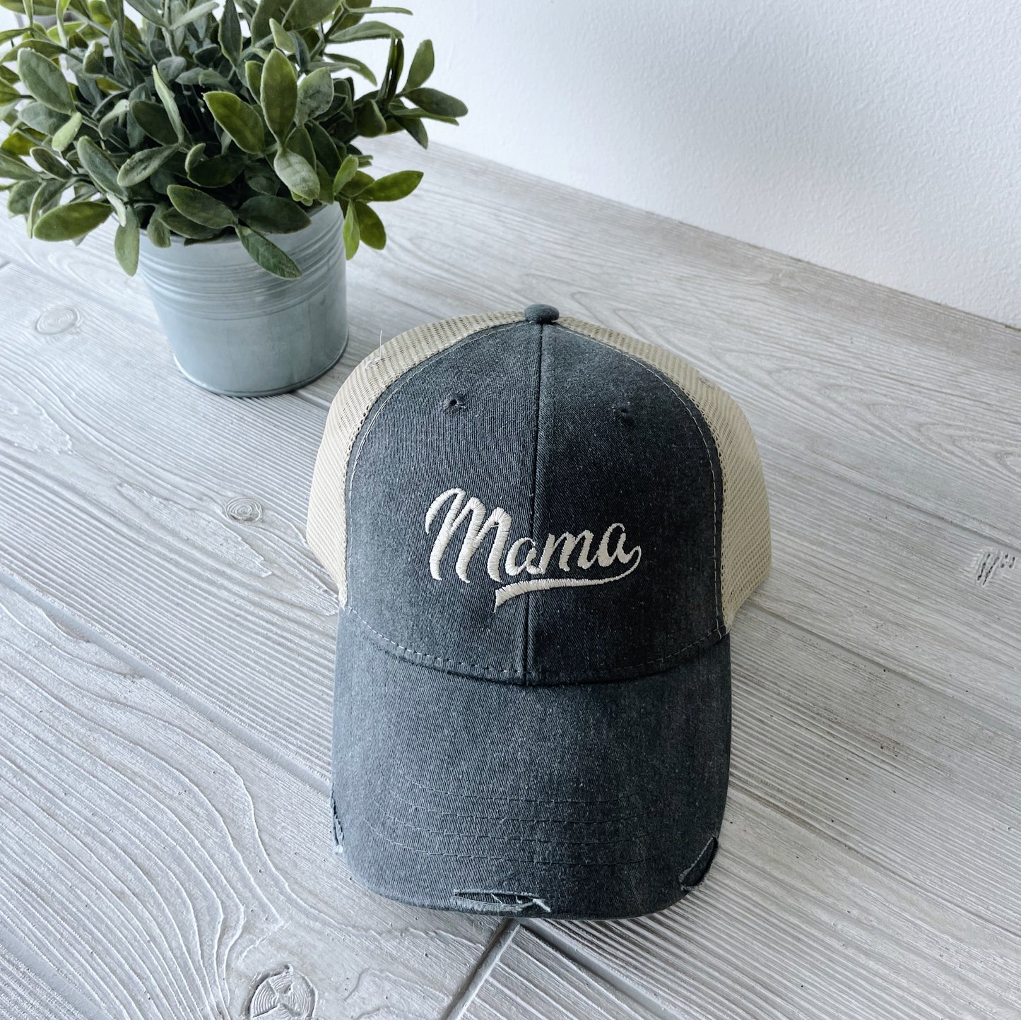 The Distressed Mama Hat • Granite features a dark gray and beige trucker style with a cotton front panel and “Mama” embroidered in white. Shown on a light wood surface beside a small potted plant, its ideal for fans of embroidered hats.